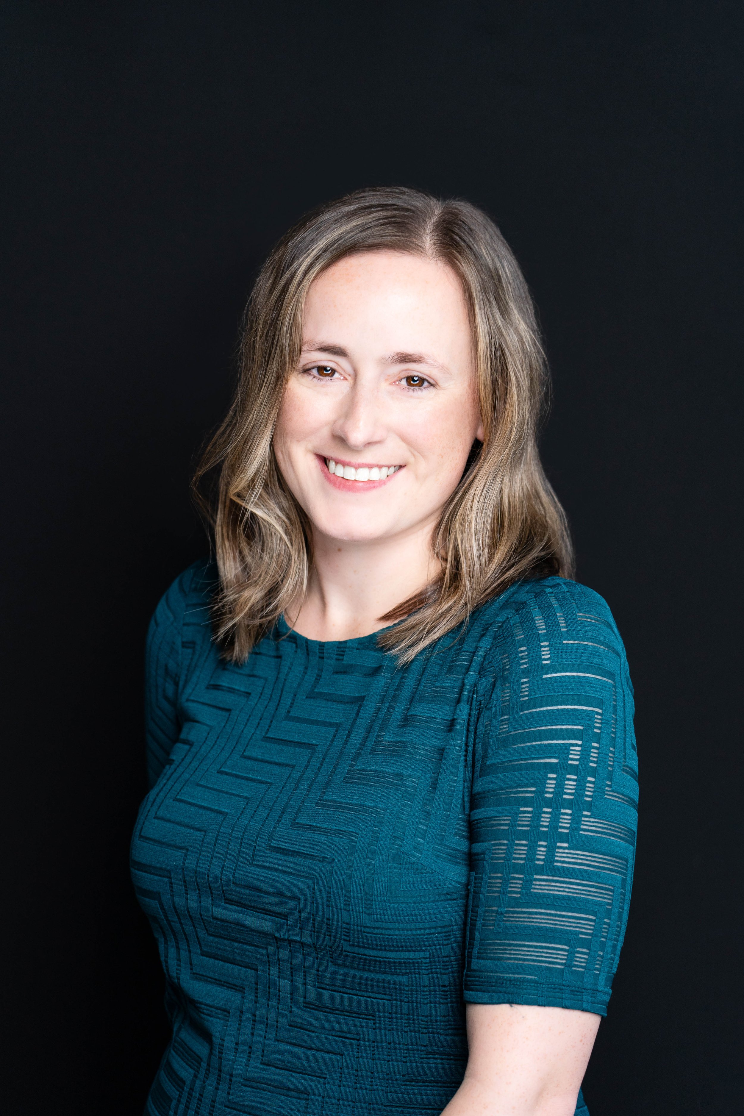 Portrait of a young woman with shoulder-length wavy brown hair, smiling, wearing a teal patterned dress, against a black background.