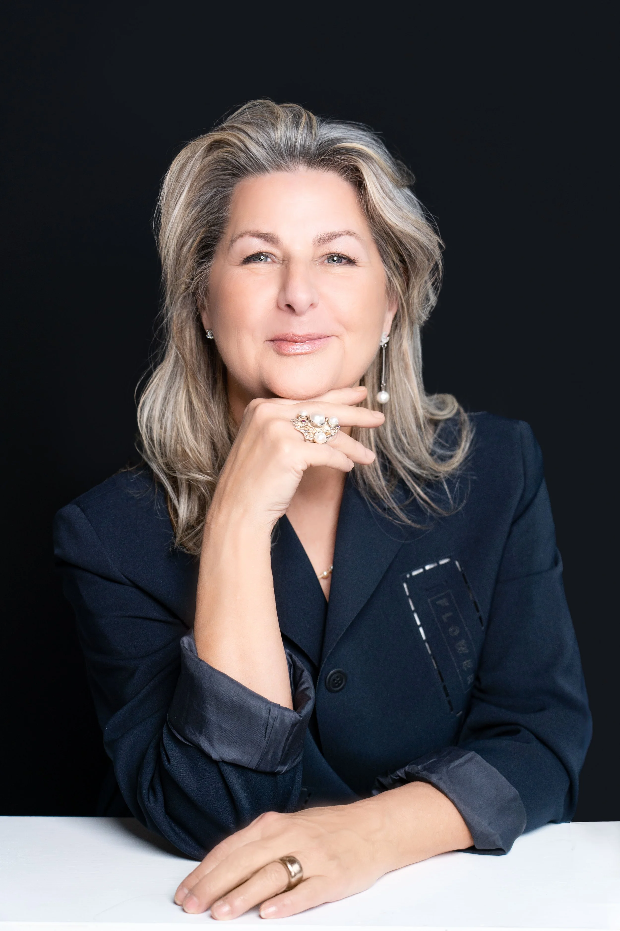 A woman with styled gray hair, wearing a navy blazer with rolled-up sleeves, sitting at a white table against a black background. She is smiling slightly, resting her chin on her right hand, and wearing earrings, a pearl ring, and a bracelet.
