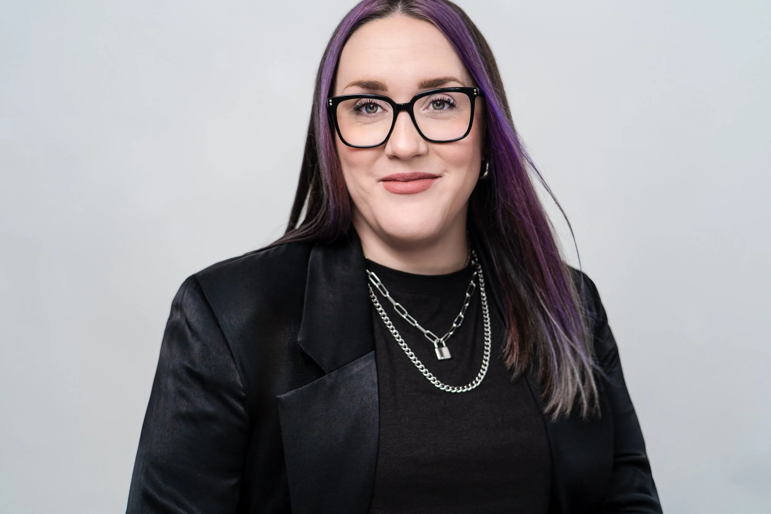 A woman with purple and black hair, wearing black glasses, black blazer, and layered silver necklaces, smiling at the camera against a plain light gray background.