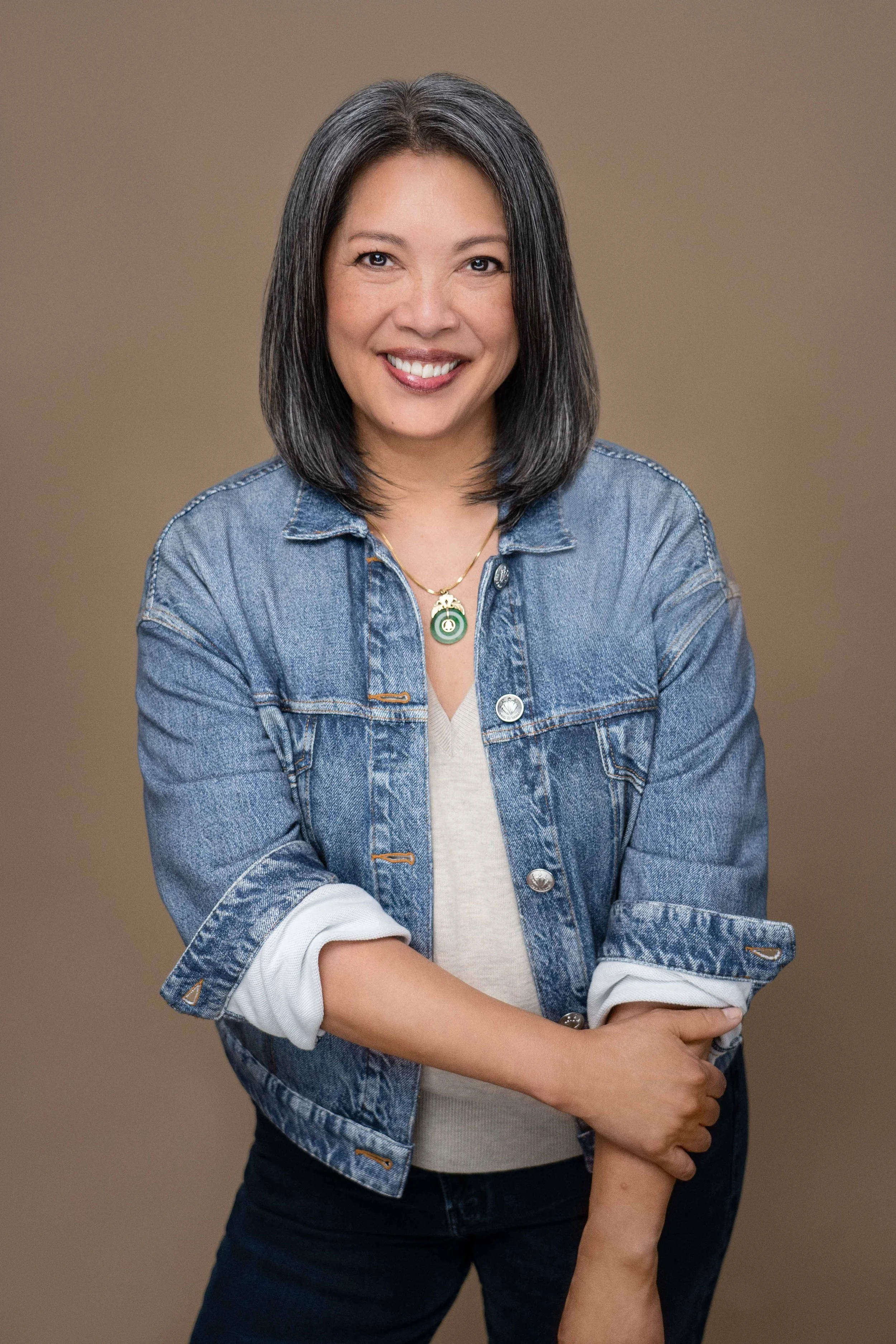 A smiling woman with shoulder-length black hair, wearing a denim jacket over a beige top, with a gold necklace, standing against a plain brown background.