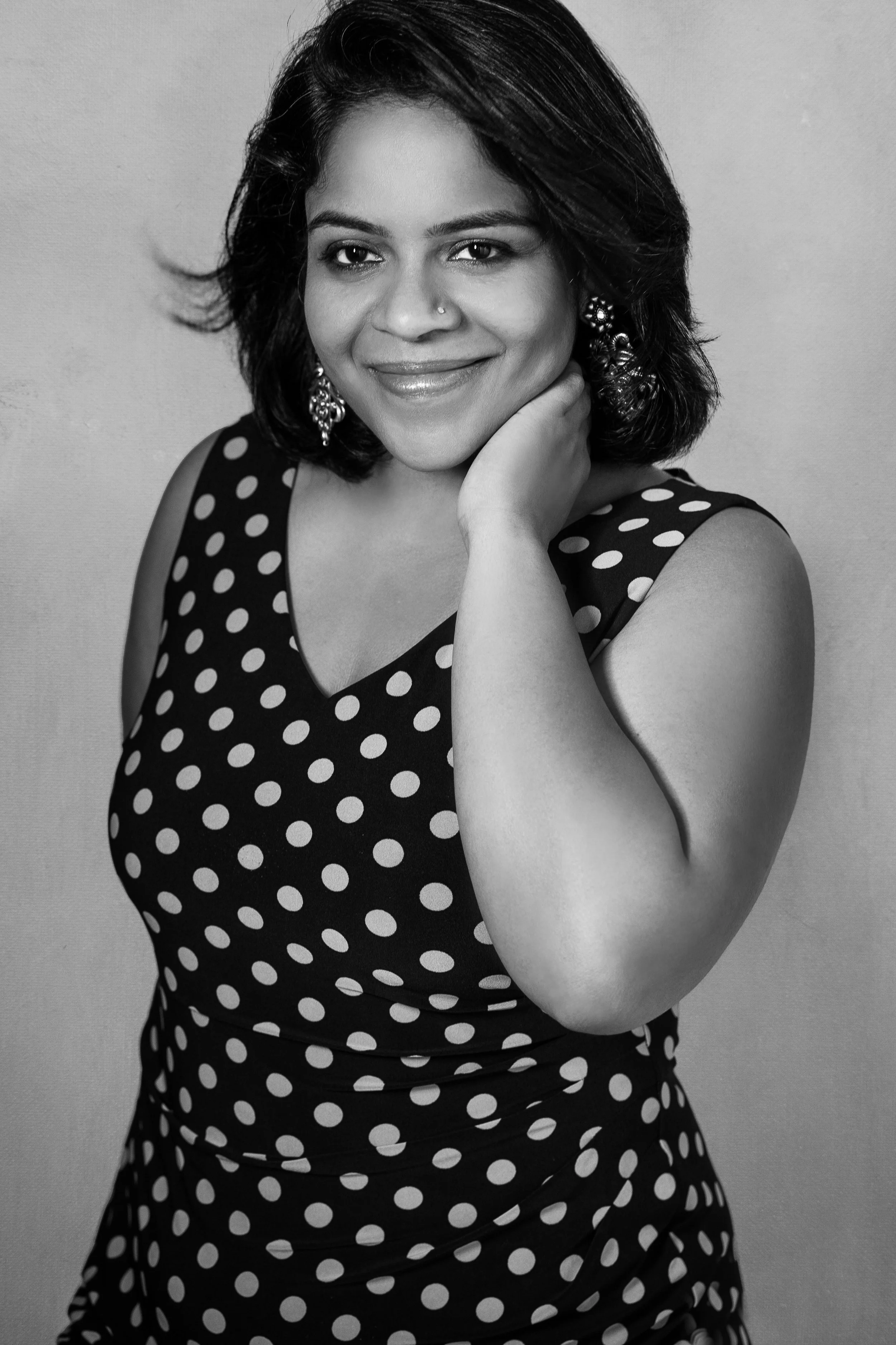 A portrait of a woman with short hair, smiling, wearing earrings, and a sleeveless polka dot dress, posed with her hand touching her face.