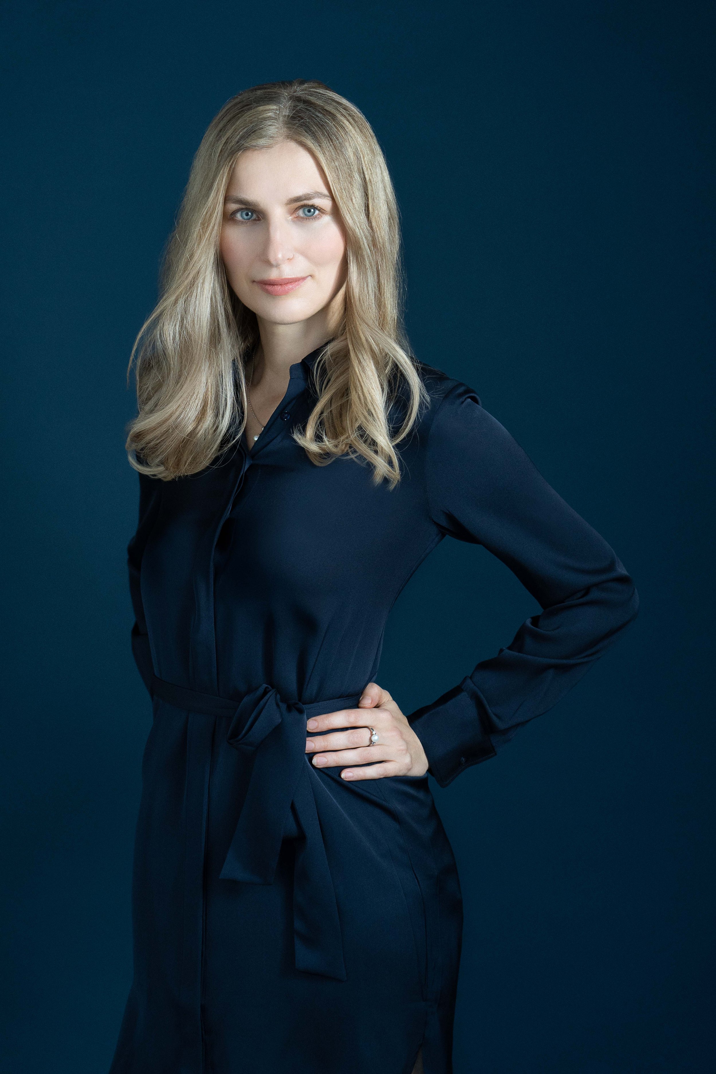 A woman with blonde hair wearing a navy blue dress standing against a dark blue background.