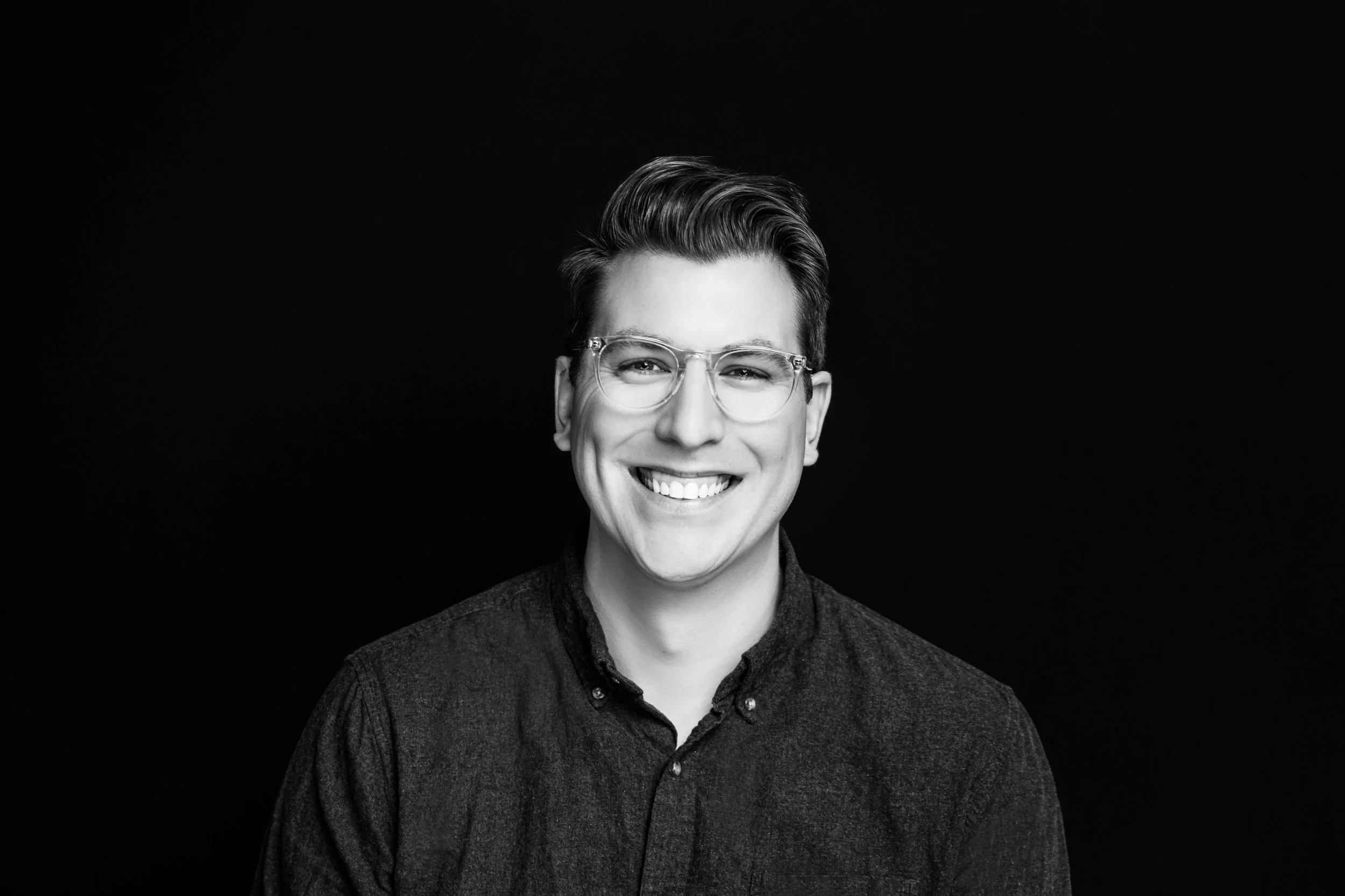 Black and white portrait of a smiling young man with glasses, short hair, and wearing a buttoned-up shirt, against a black background.