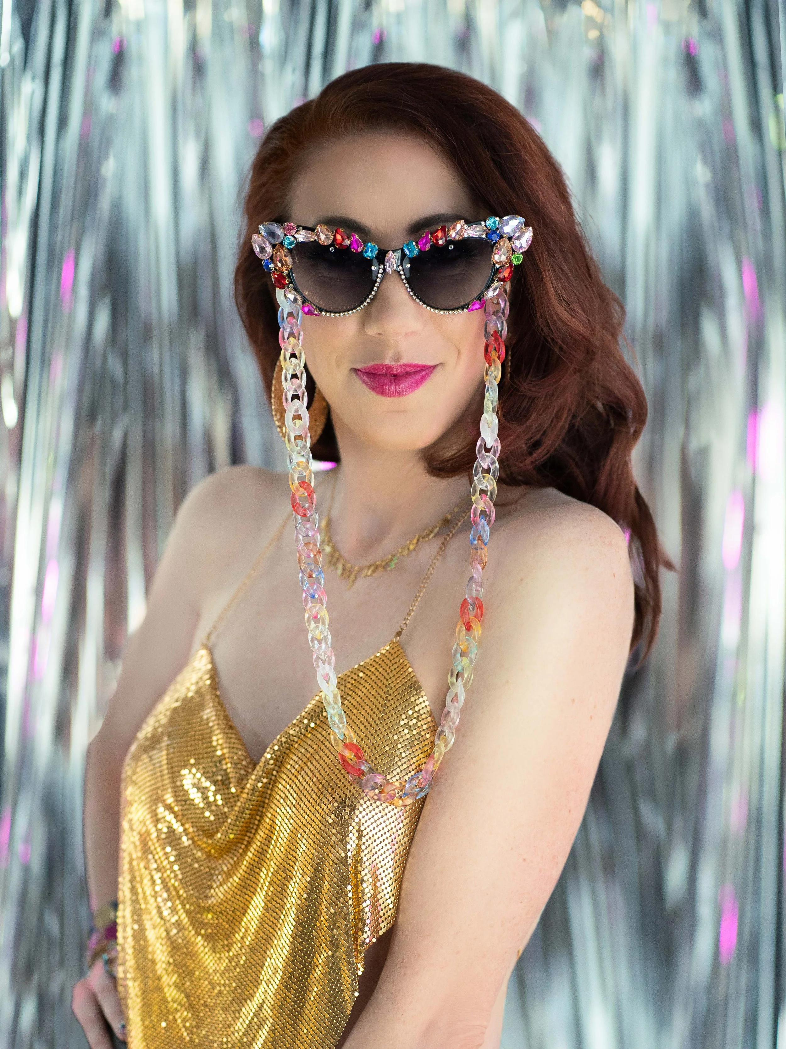 A woman with red hair wearing black sunglasses adorned with colorful jewels, a gold sequin top, and colorful chain necklace, standing in front of shiny silver decorations.