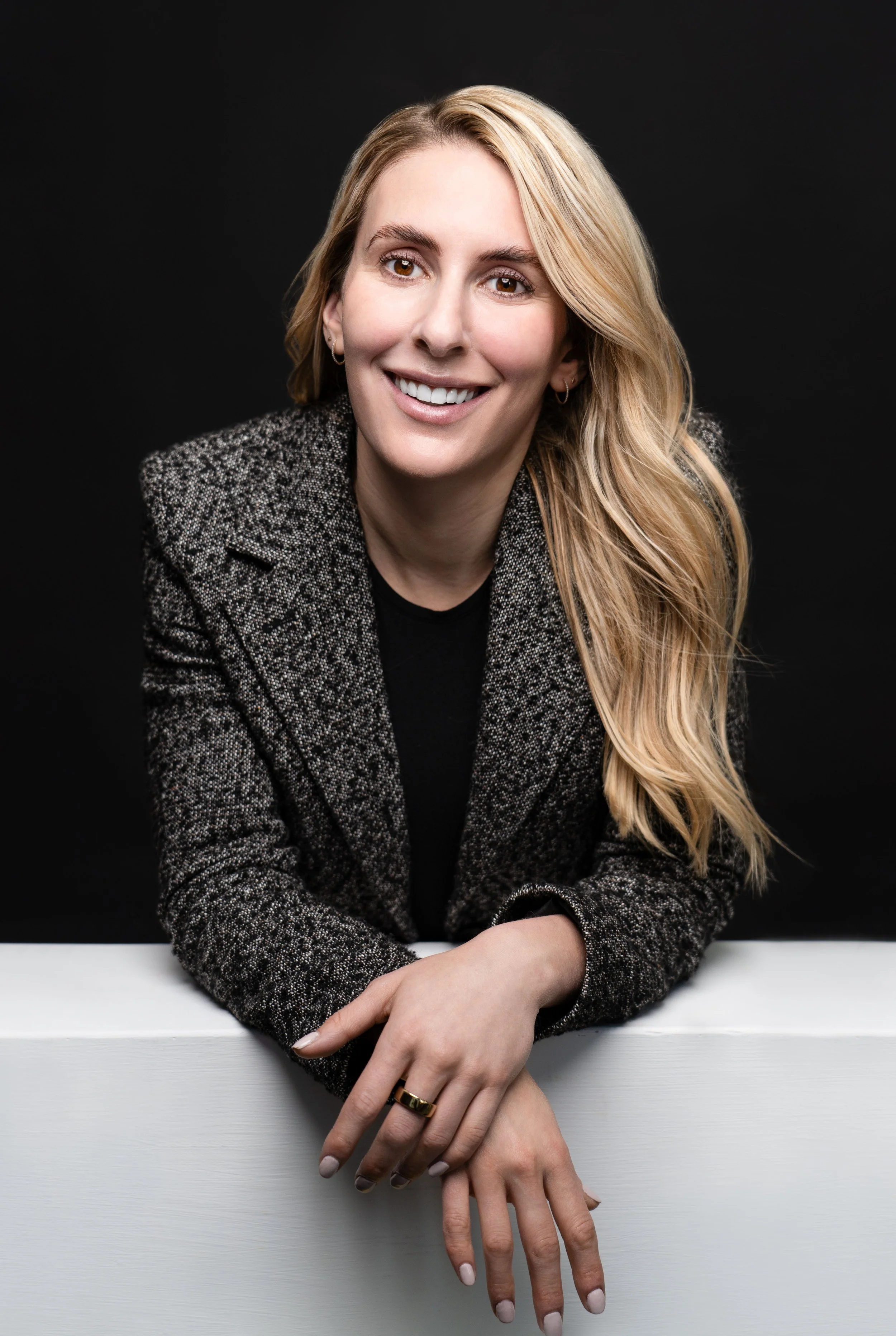 Portrait of a smiling woman with long blonde hair, wearing a patterned blazer, leaning on a white surface against a black background.