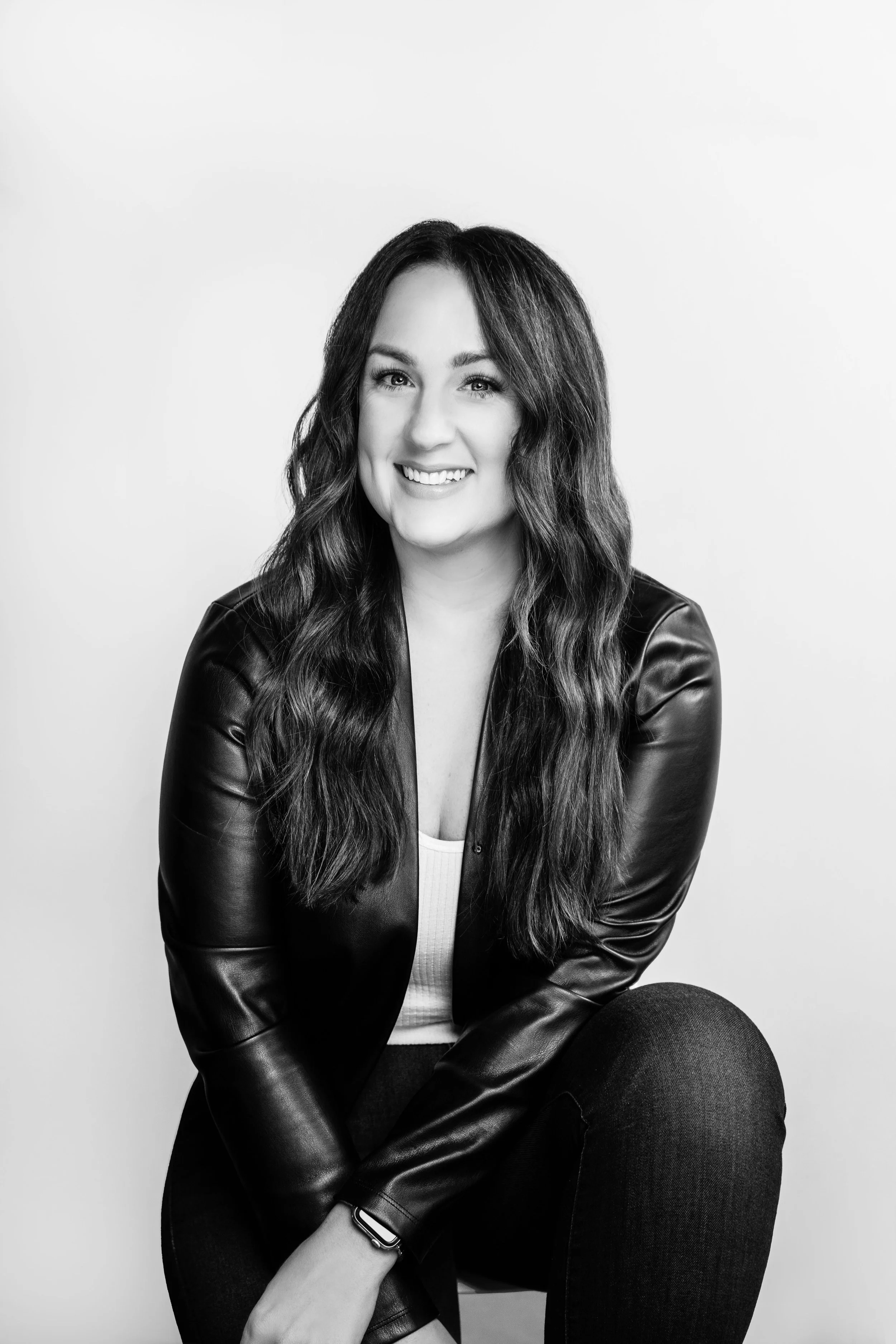 A portrait of a woman with long wavy dark hair, smiling, wearing a leather jacket over a light top, sitting against a plain light background.