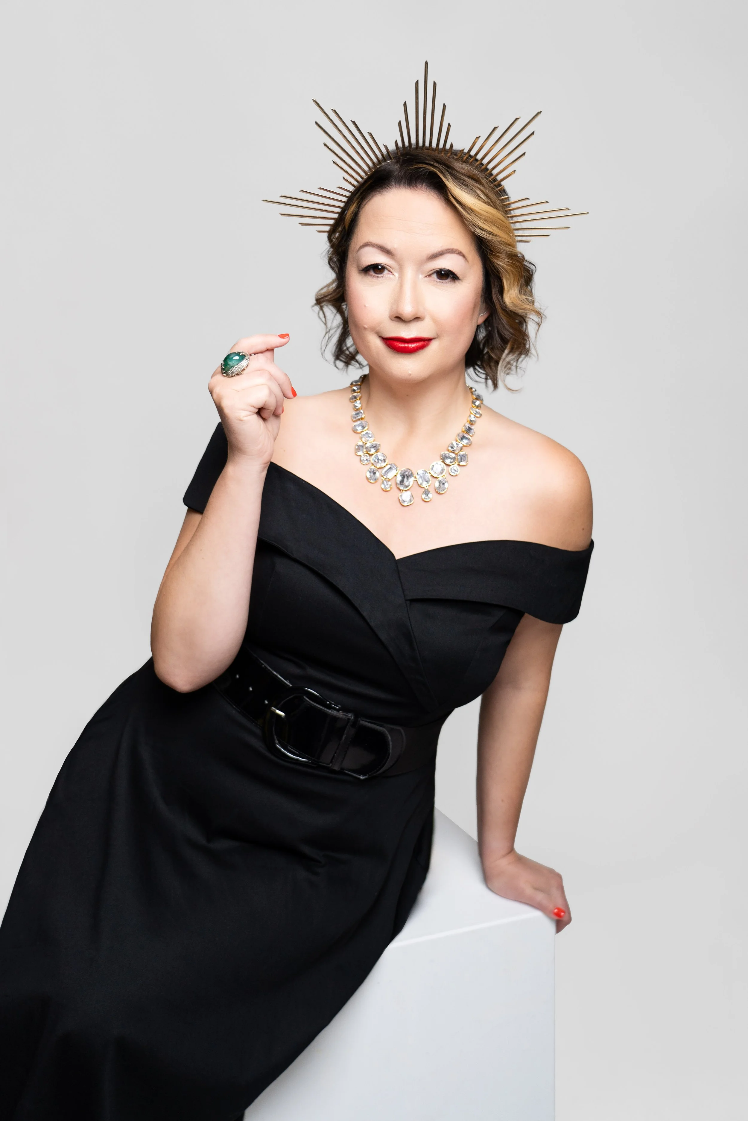 Woman wearing a black off-shoulder dress, large diamond necklace, green ring, red lipstick, and a bronze sunburst headpiece, posing against a plain gray background.