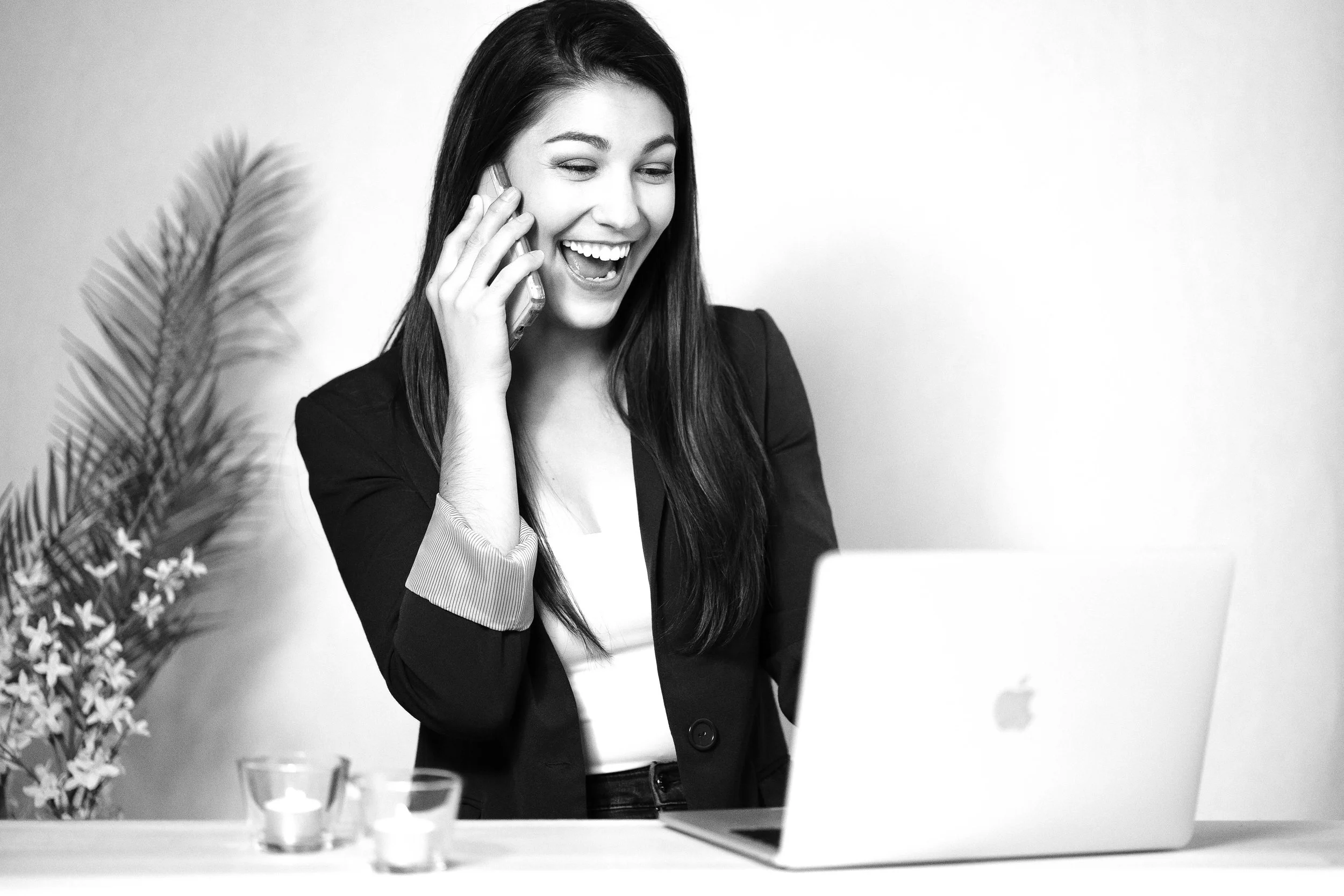 A woman with long dark hair smiling and talking on her cellphone while looking at her open laptop on a desk with candles and a plant in the background.