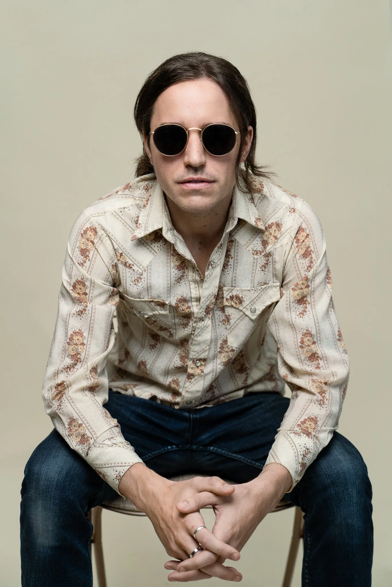 A man with dark hair, wearing round black sunglasses, a beige floral-patterned shirt, and dark jeans, is sitting on a chair against a plain light-colored background.