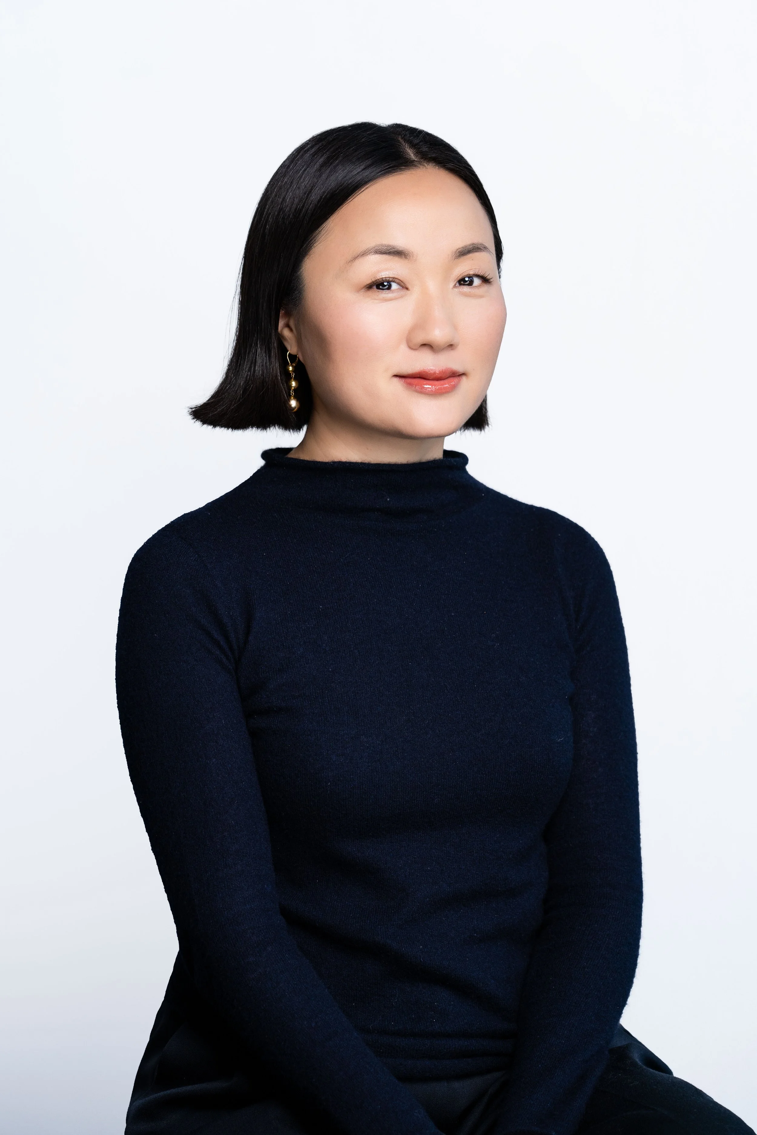 A woman with straight dark hair, wearing a black turtleneck sweater and gold earrings, posing against a plain white background.