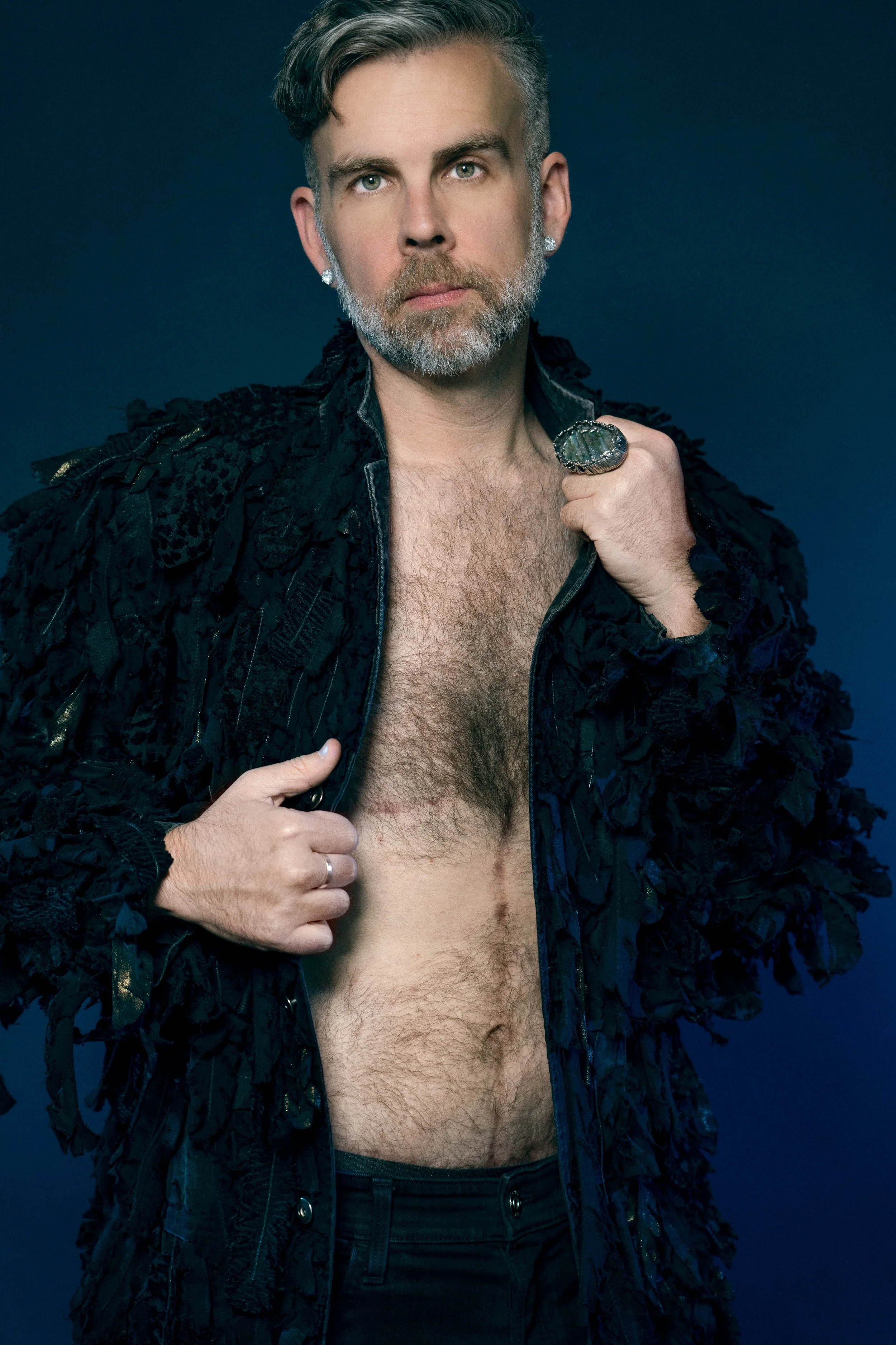 Man with styled hair and beard posing against a dark background, partially unzipping a textured black jacket to reveal chest hair, wearing jewelry including large rings and earrings.