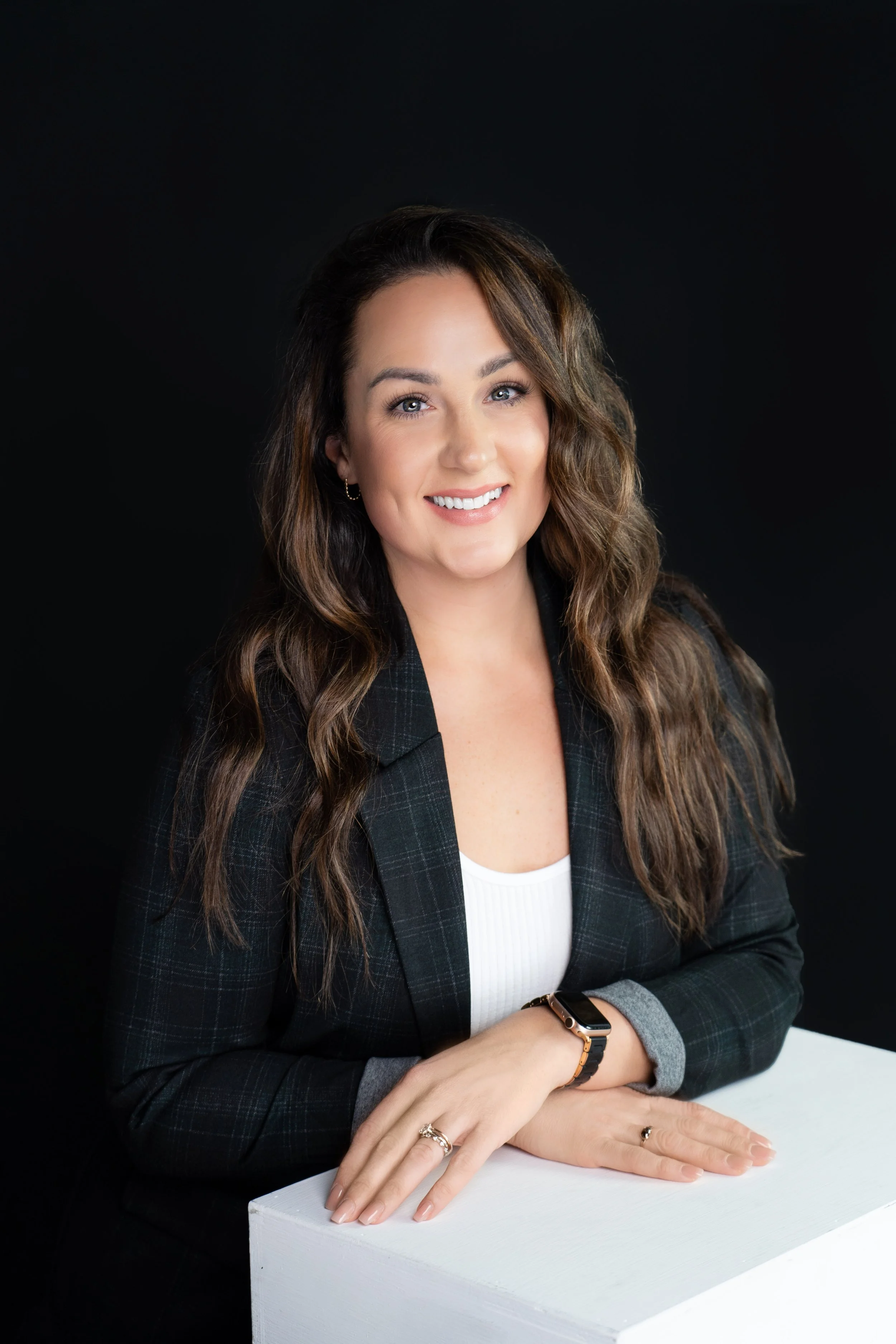 A woman with long wavy brown hair and light skin, wearing a dark plaid blazer, white top, and a smartwatch, sitting behind a white table against a black background, smiling at the camera.