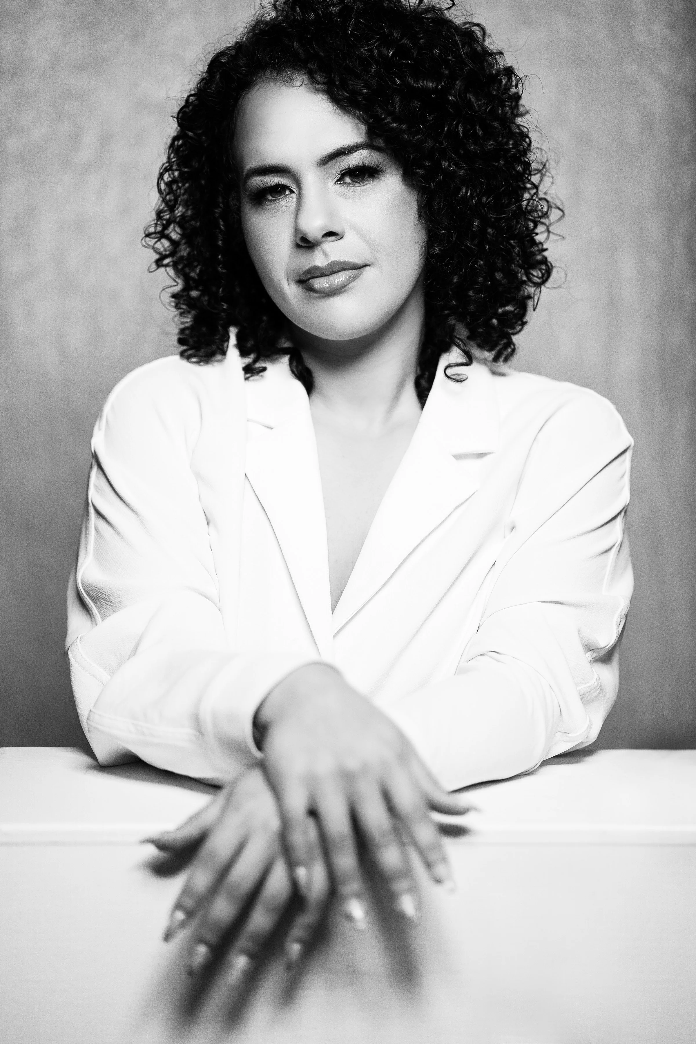 Black and white portrait of a woman with curly dark hair, wearing a white blazer, sitting at a table, with her hands resting on the surface, looking directly at the camera with a confident expression.
