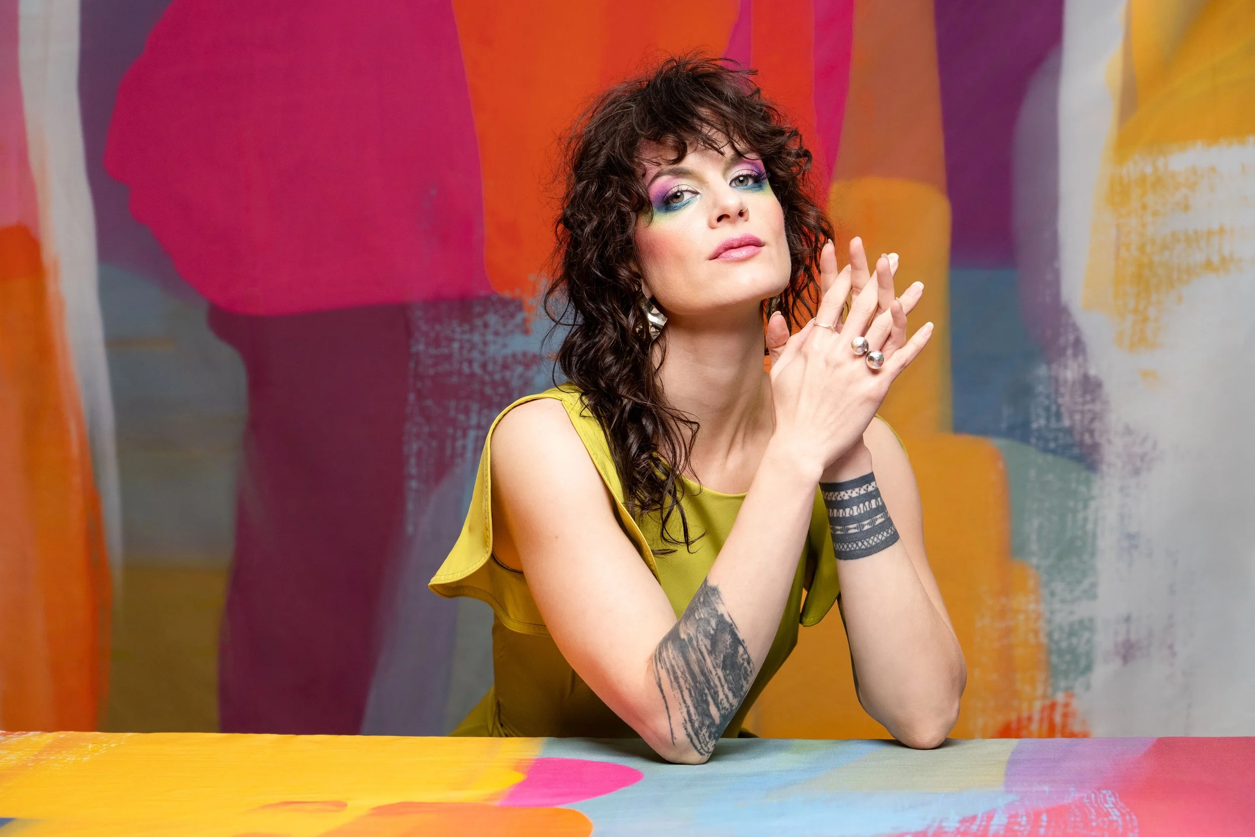A woman with curly dark hair and colorful makeup poses with hands clasped together in front of a vibrant, abstract, multicolored background, wearing a yellow sleeveless top and multiple rings and tattoos.