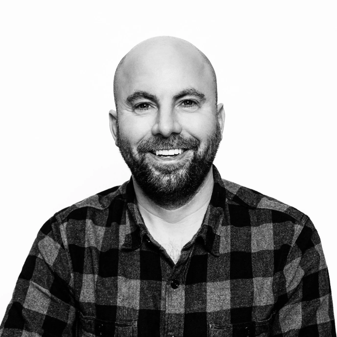 Black and white portrait of a smiling man with a beard and bald head, wearing a plaid shirt.