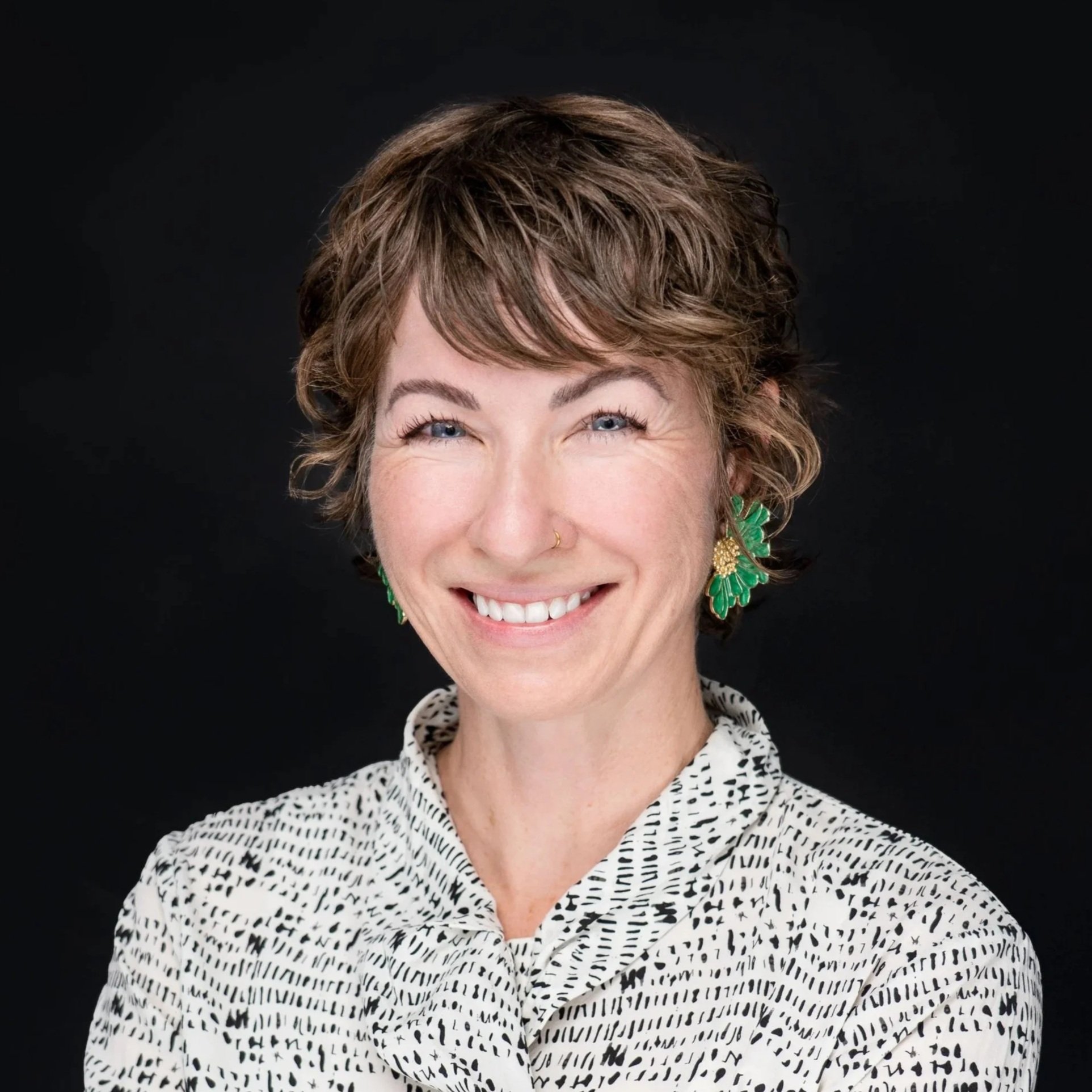 A headshot woman with short brown hair green earrings and a white blouse smiling against a black background