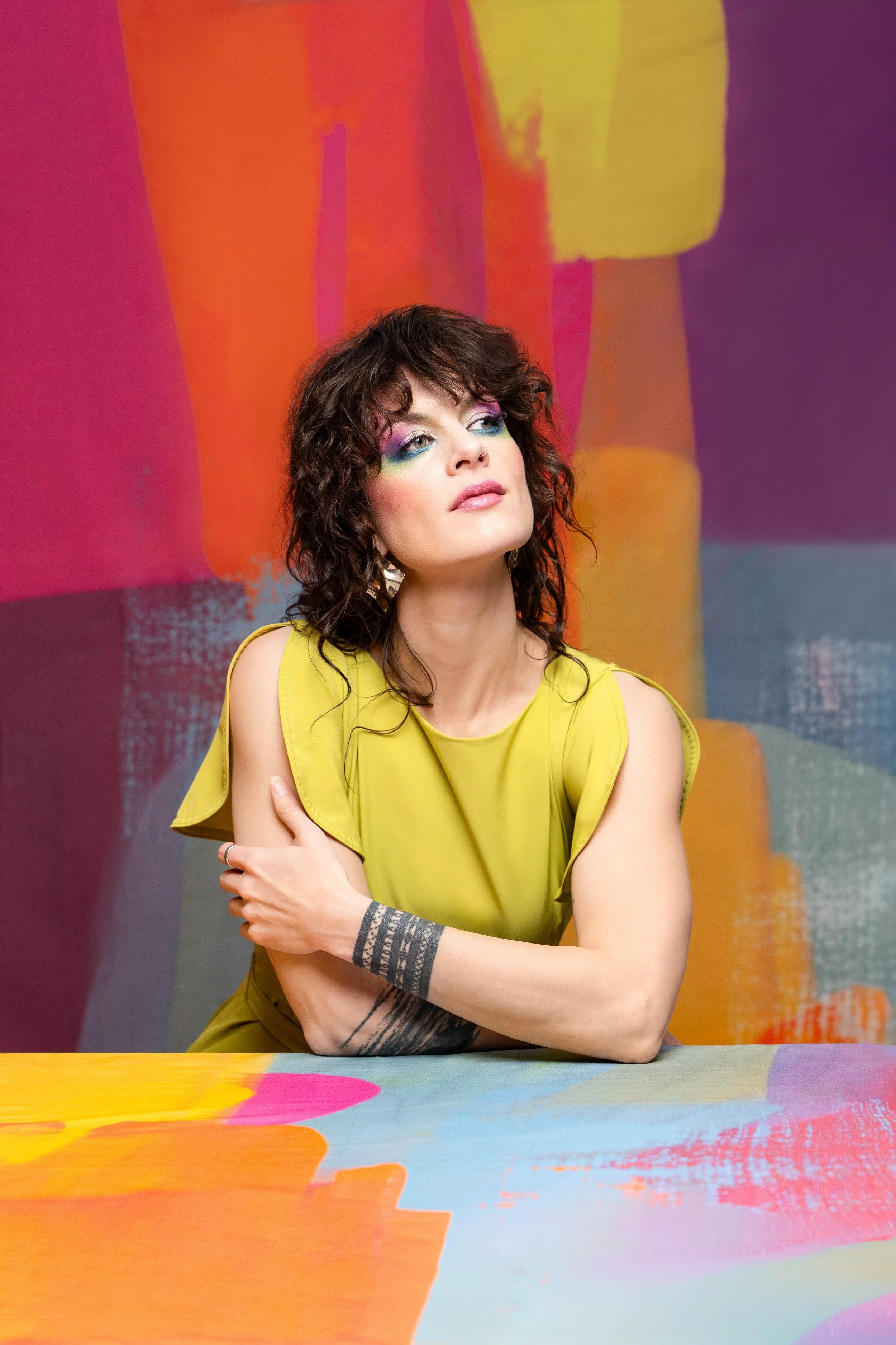 A woman with curly dark hair and colorful makeup, wearing a yellow sleeveless dress, sitting at a painted table with a multicolored abstract background.