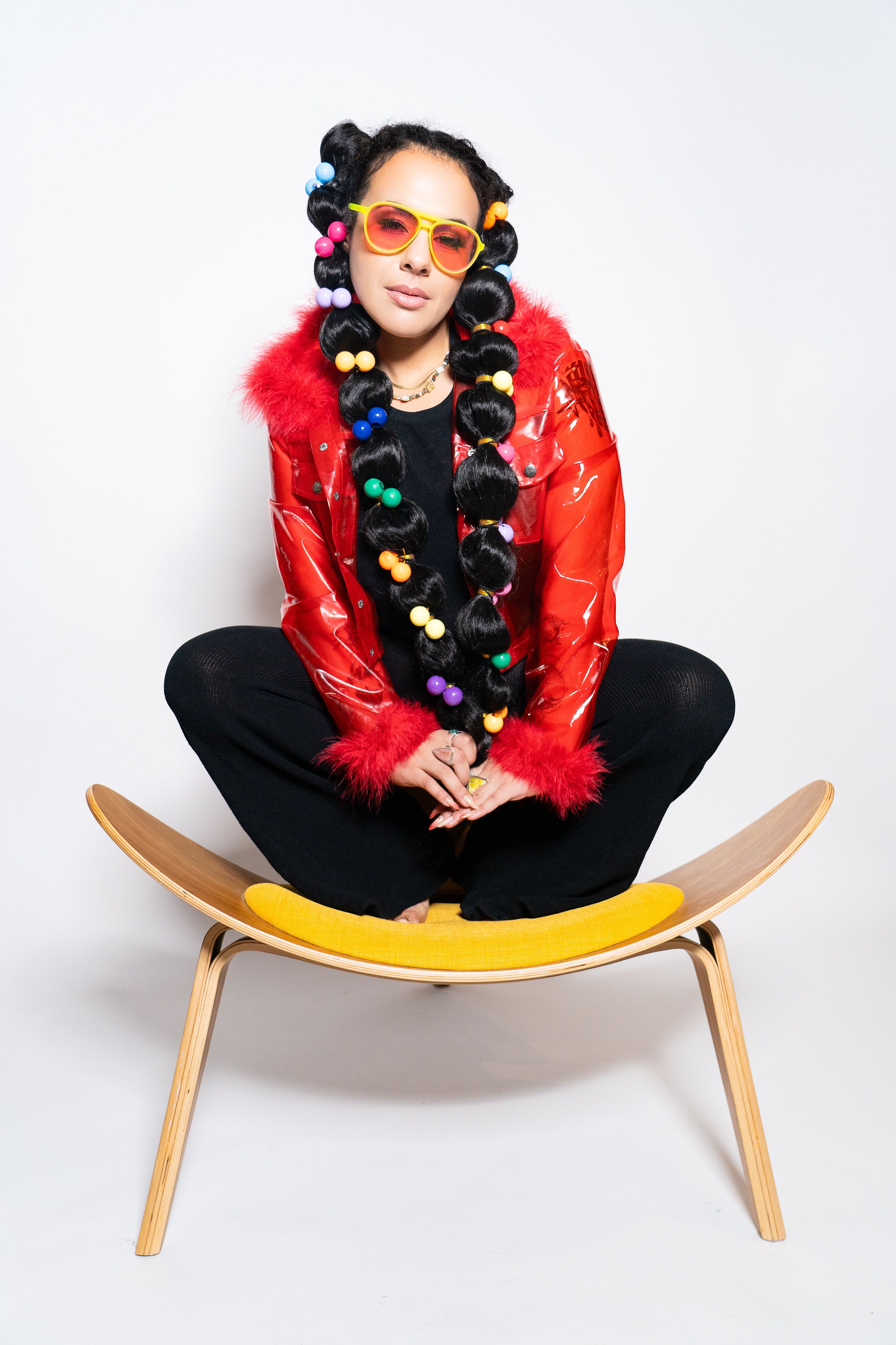 A woman sitting cross-legged on a yellow cushioned wooden chair against a plain white background. She has long, black hair styled into large buns with colorful hair beads and wears large yellow sunglasses, a shiny red jacket with red fur trim, black 