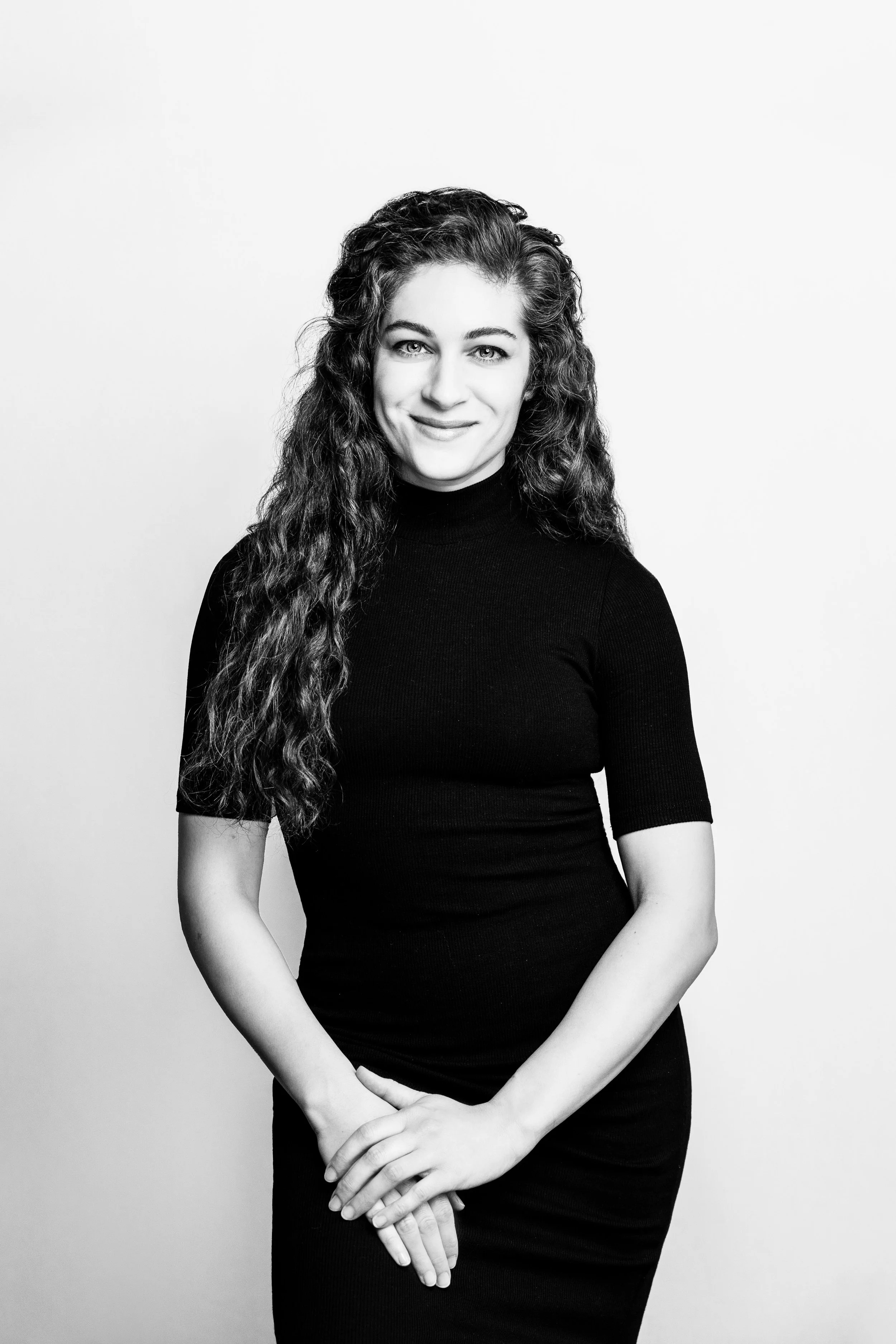 Black and white portrait of a woman with long, curly hair wearing a black turtleneck and dress, smiling softly and standing against a plain light background.