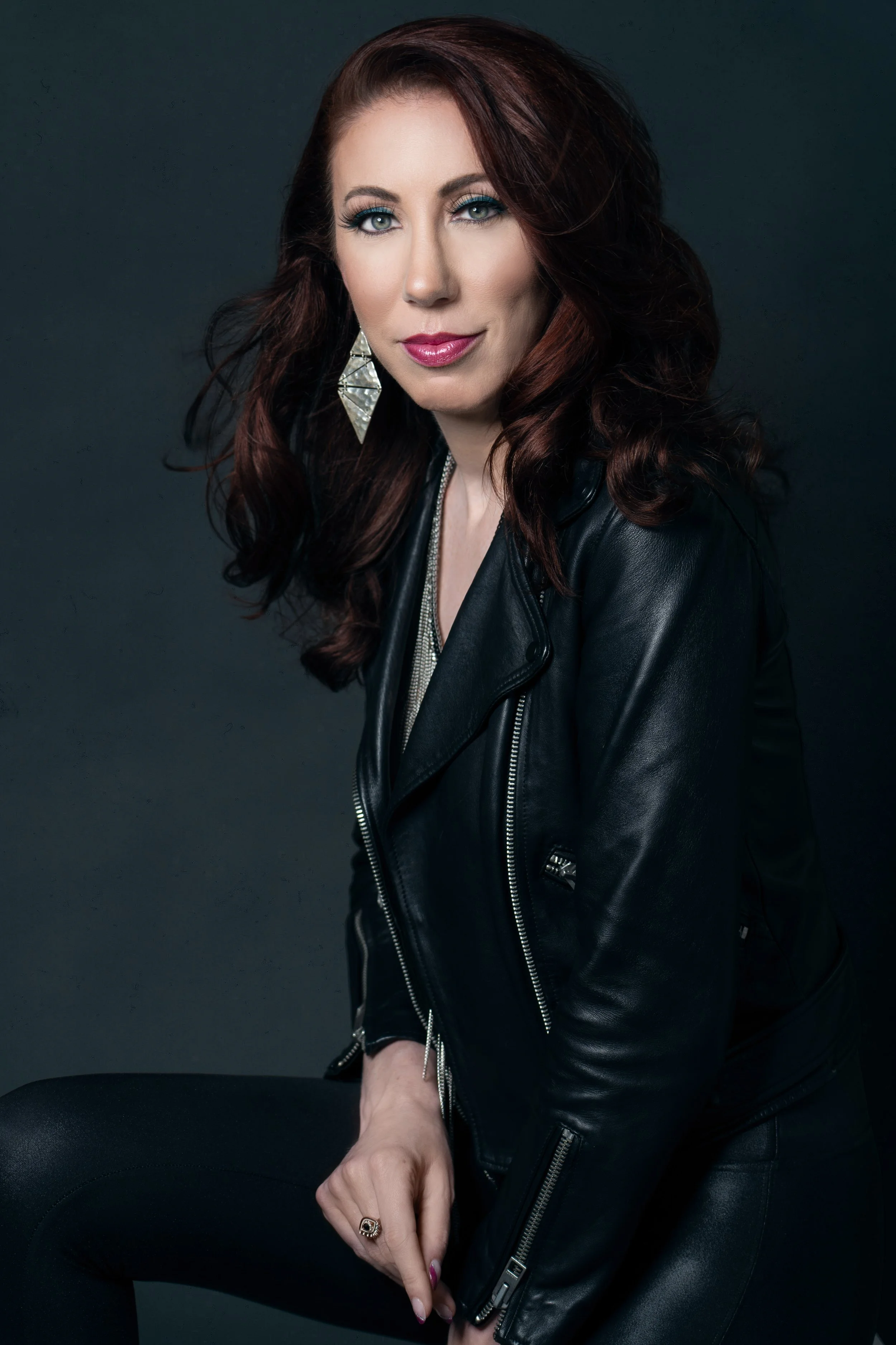 A woman with shoulder-length red hair, wearing a black leather jacket and large silver earrings, sitting against a dark background.