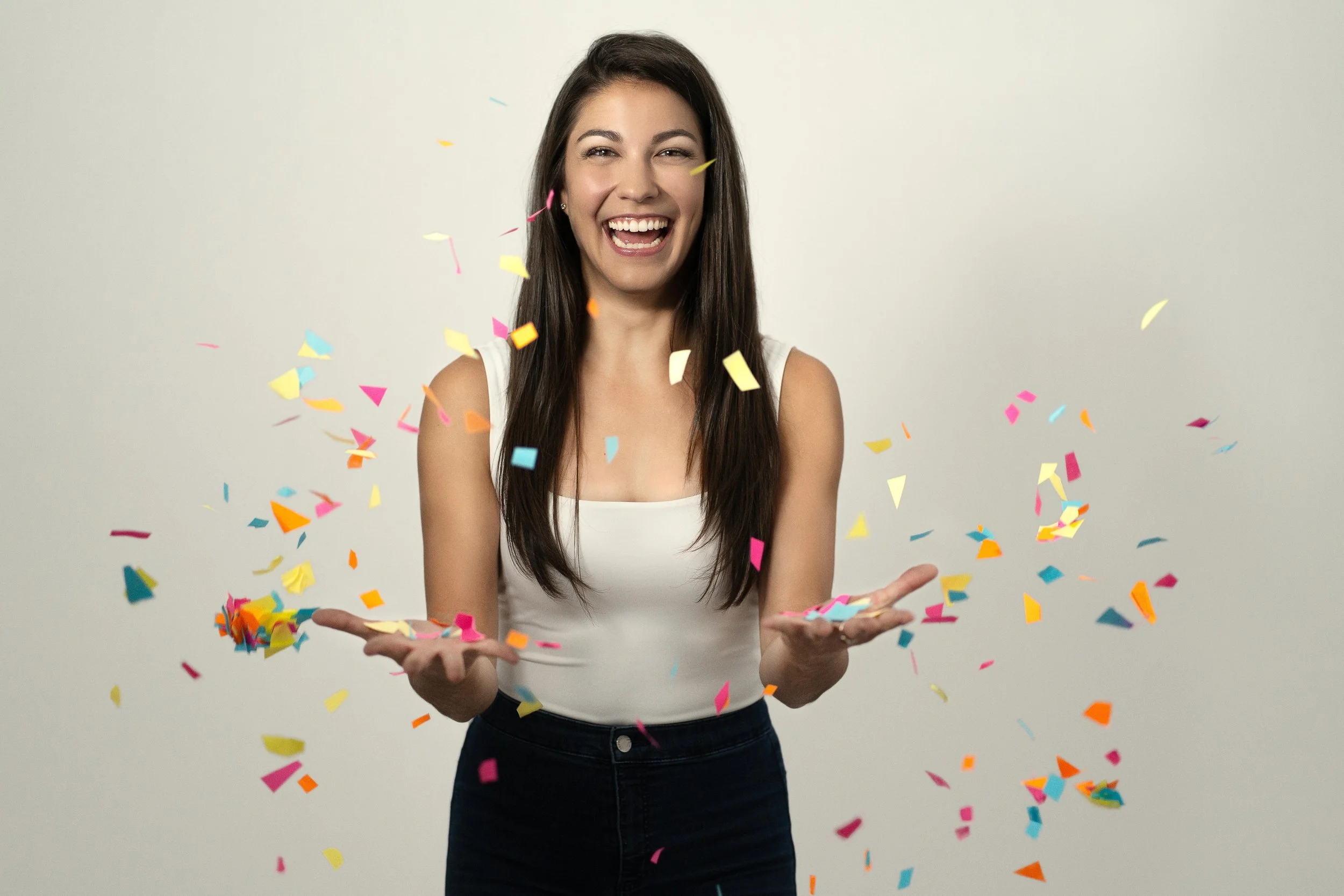 A woman with long brown hair wearing a white sleeveless top celebrating with colorful confetti falling around her and smiling broadly.