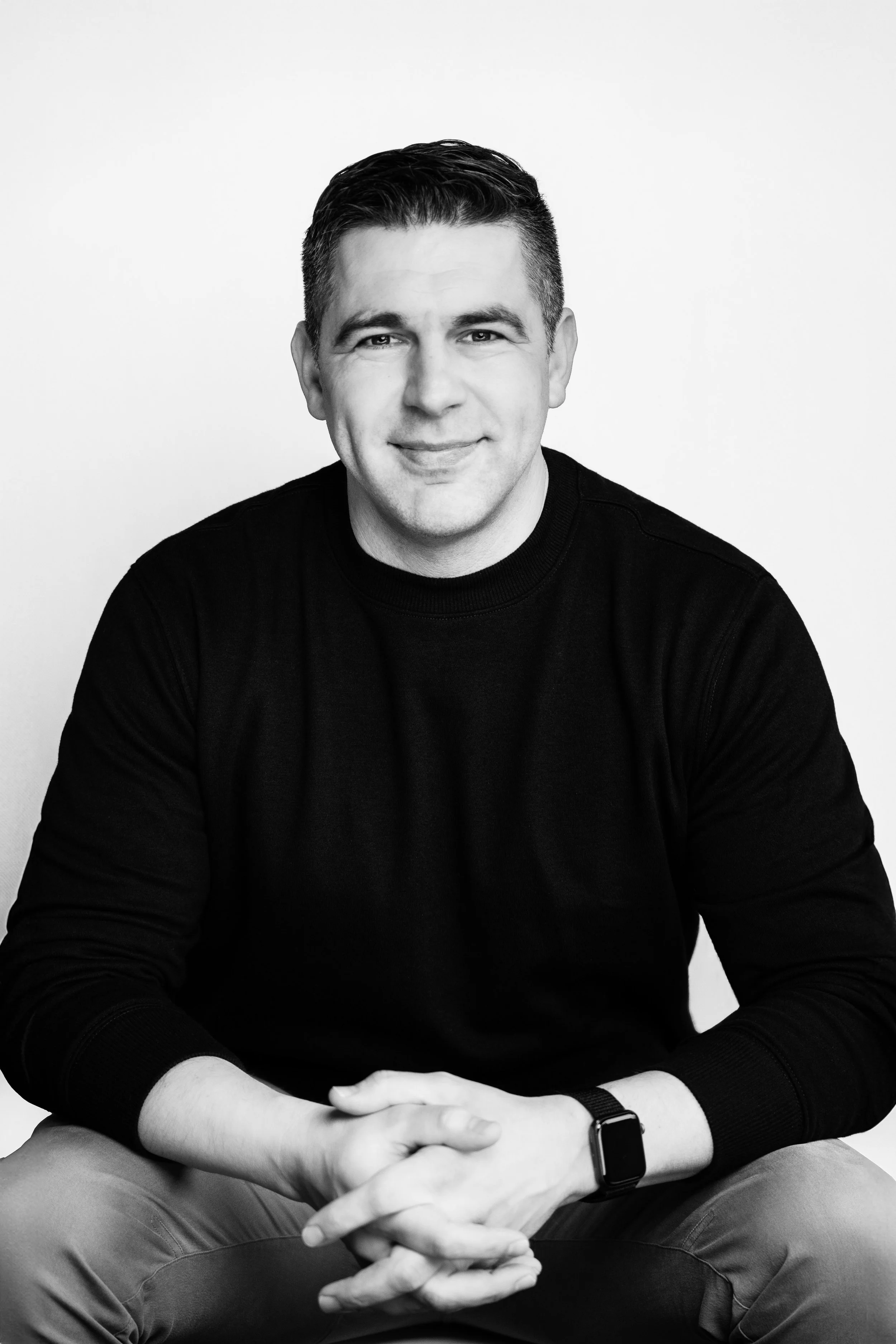 Black and white portrait of a smiling man wearing a black sweater and watch, sitting with hands clasped, against a plain white background.