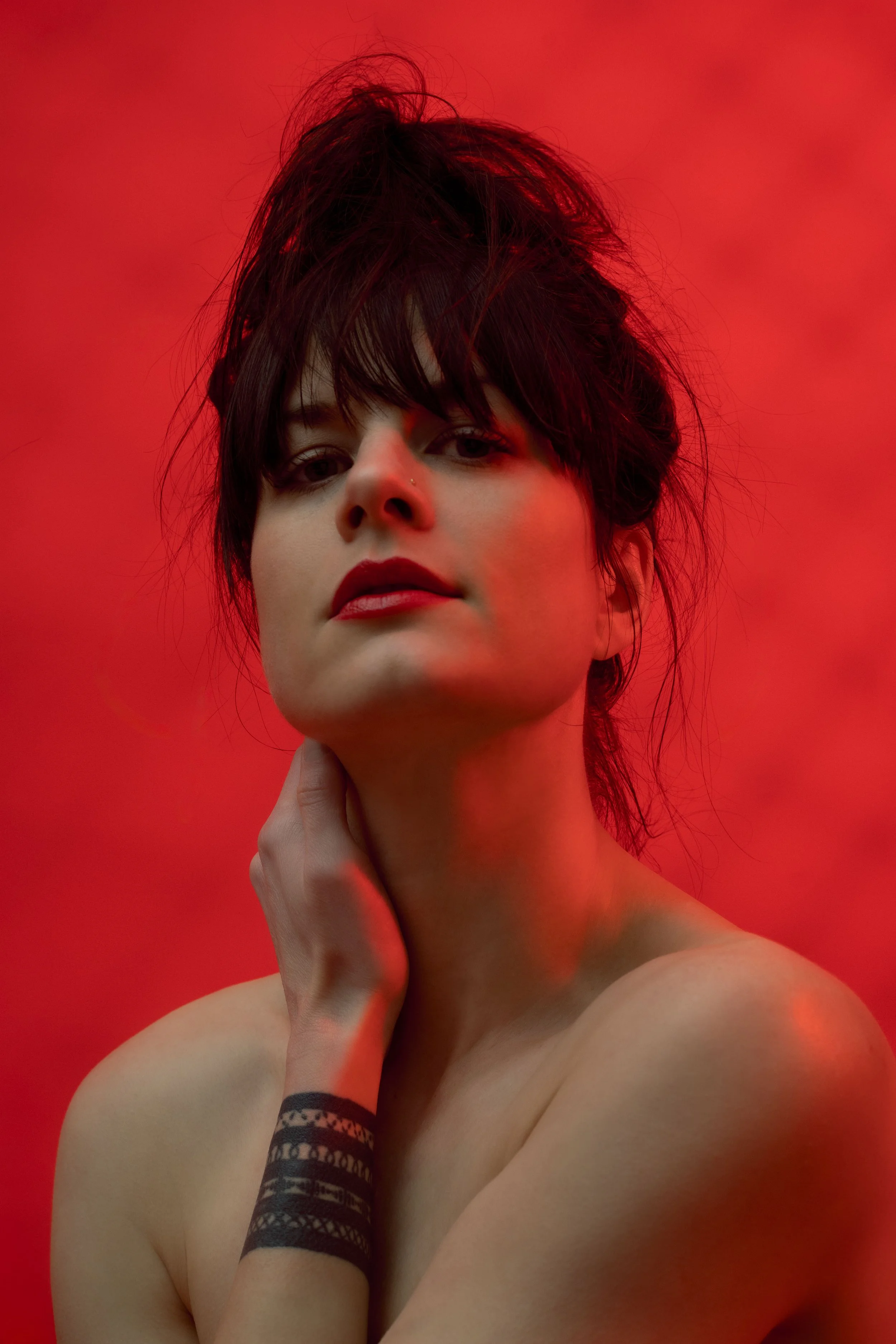 A woman with short, dark, messy hair posing against a red backdrop, looking directly at the camera with her hand touching her neck.
