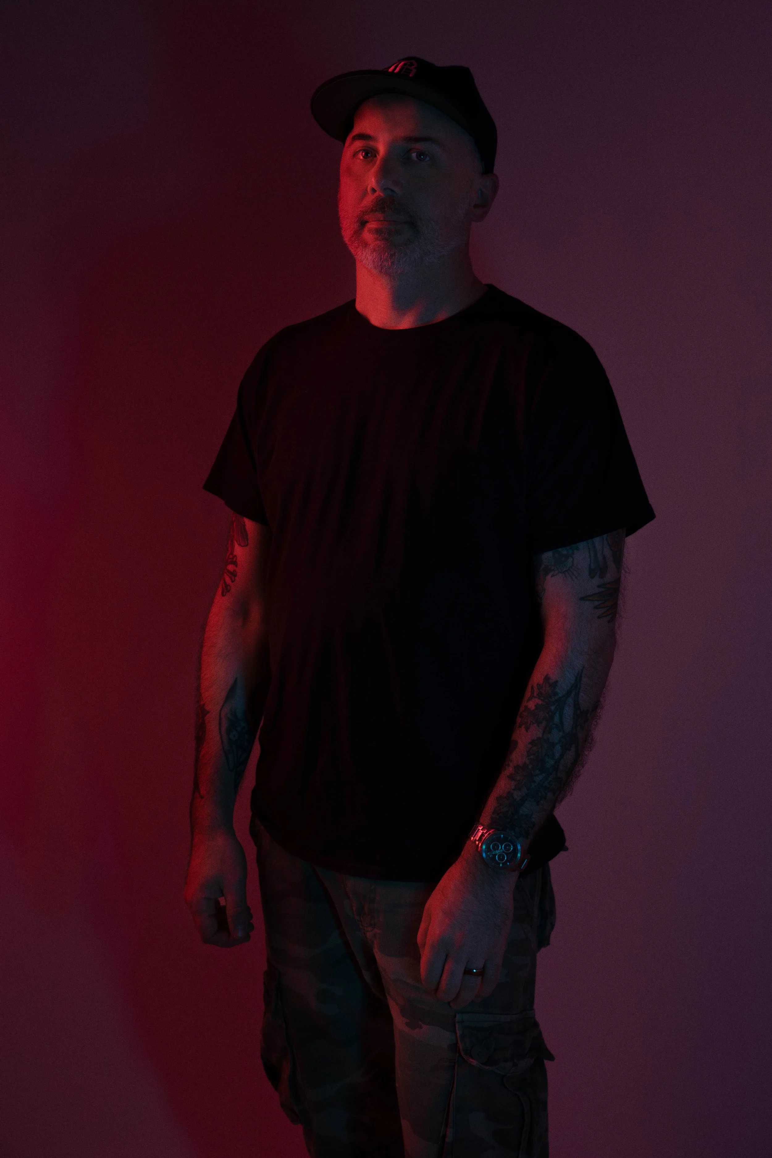 A man with tattoos on his arms, wearing a black T-shirt, camouflage pants, a black cap, and a wristwatch, standing against a dark background with red and purple lighting.