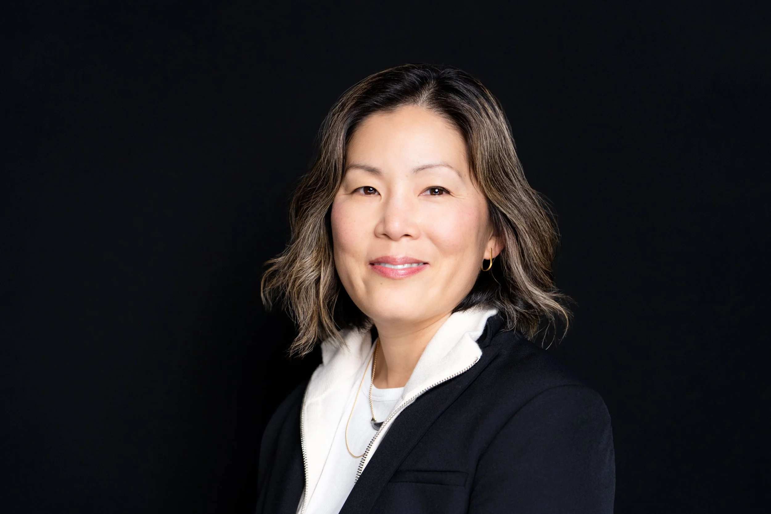 Portrait of a woman with shoulder-length wavy hair, wearing a black blazer and white hoodie, smiling softly against a black background.
