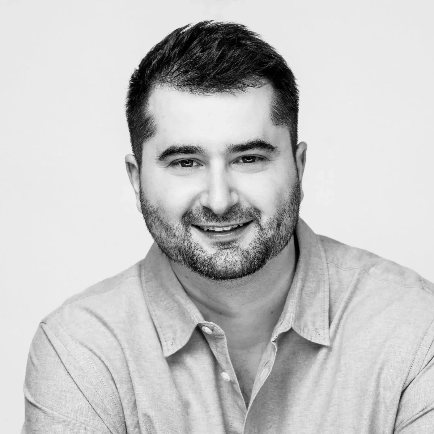 Black and white headshot of a smiling young man with short dark hair, a beard, wearing a button-up shirt.
