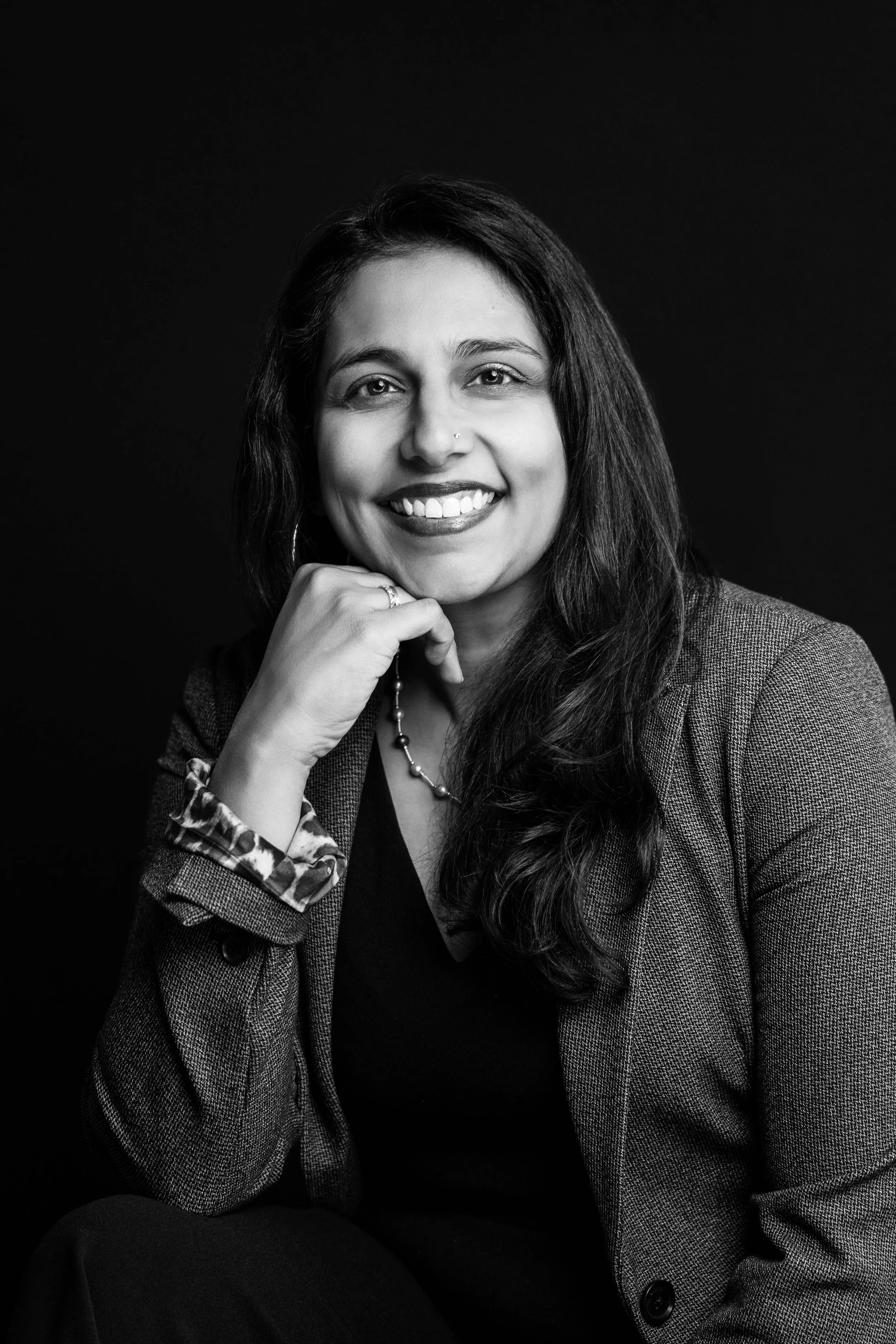 A black and white portrait of a woman with long dark hair, smiling, wearing a blazer, with her hand resting on her chin, against a plain dark background.