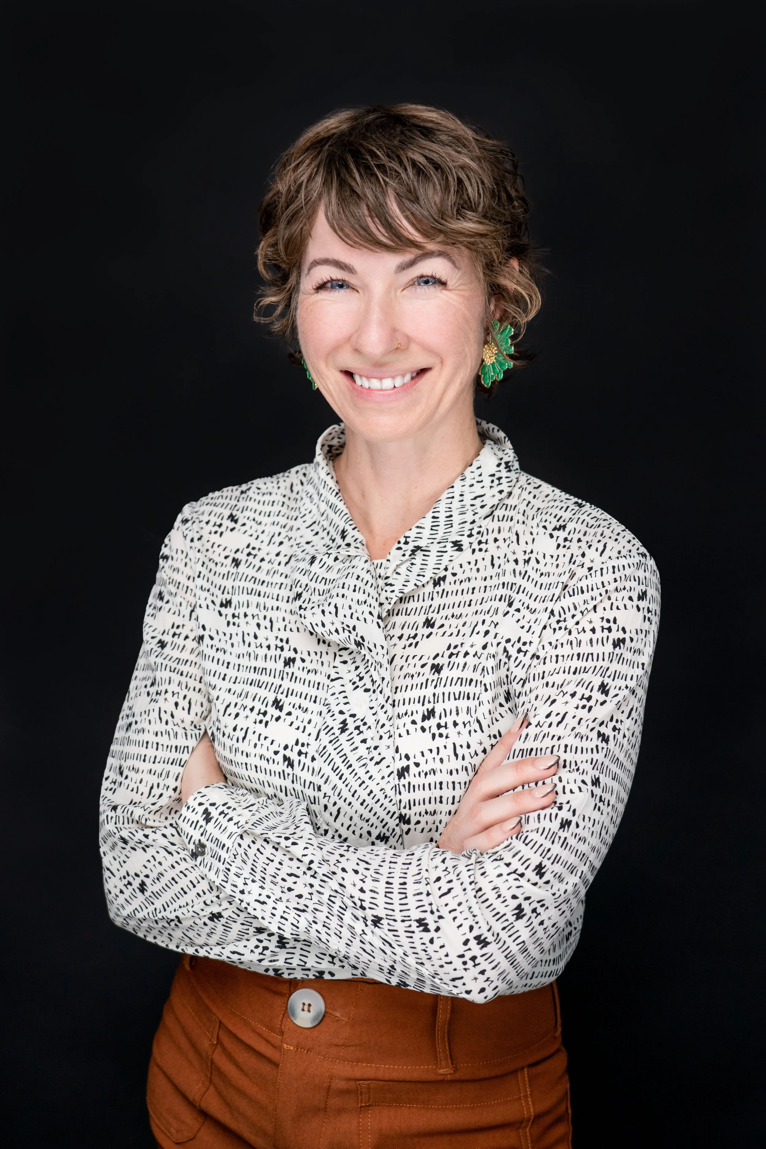 A woman with short, curly brown hair wearing a white and black patterned blouse, brown pants, and green leaf-shaped earrings, posing with her arms crossed against a black background.