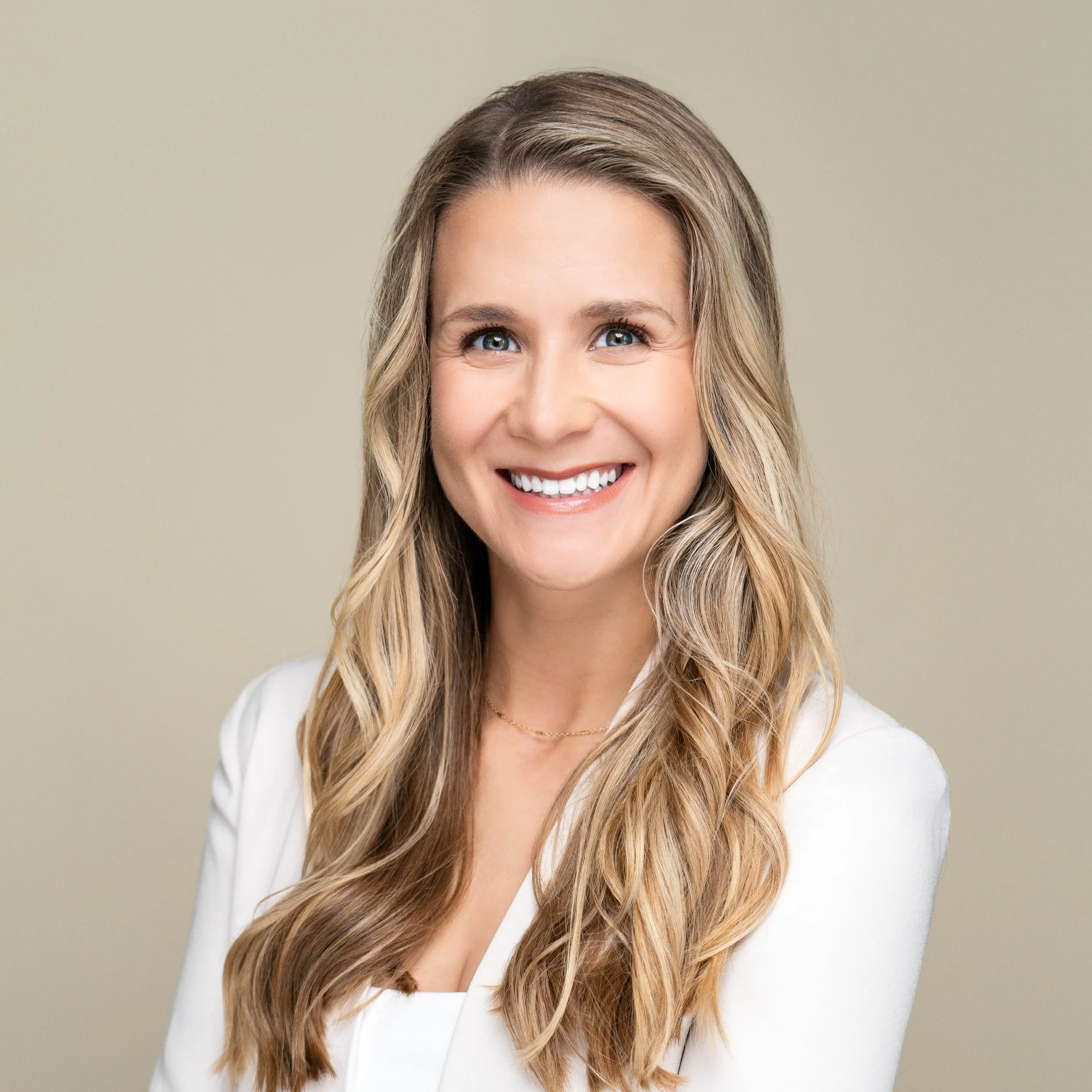 A woman with long wavy blonde hair smiling in a white blazer against a neutral background.