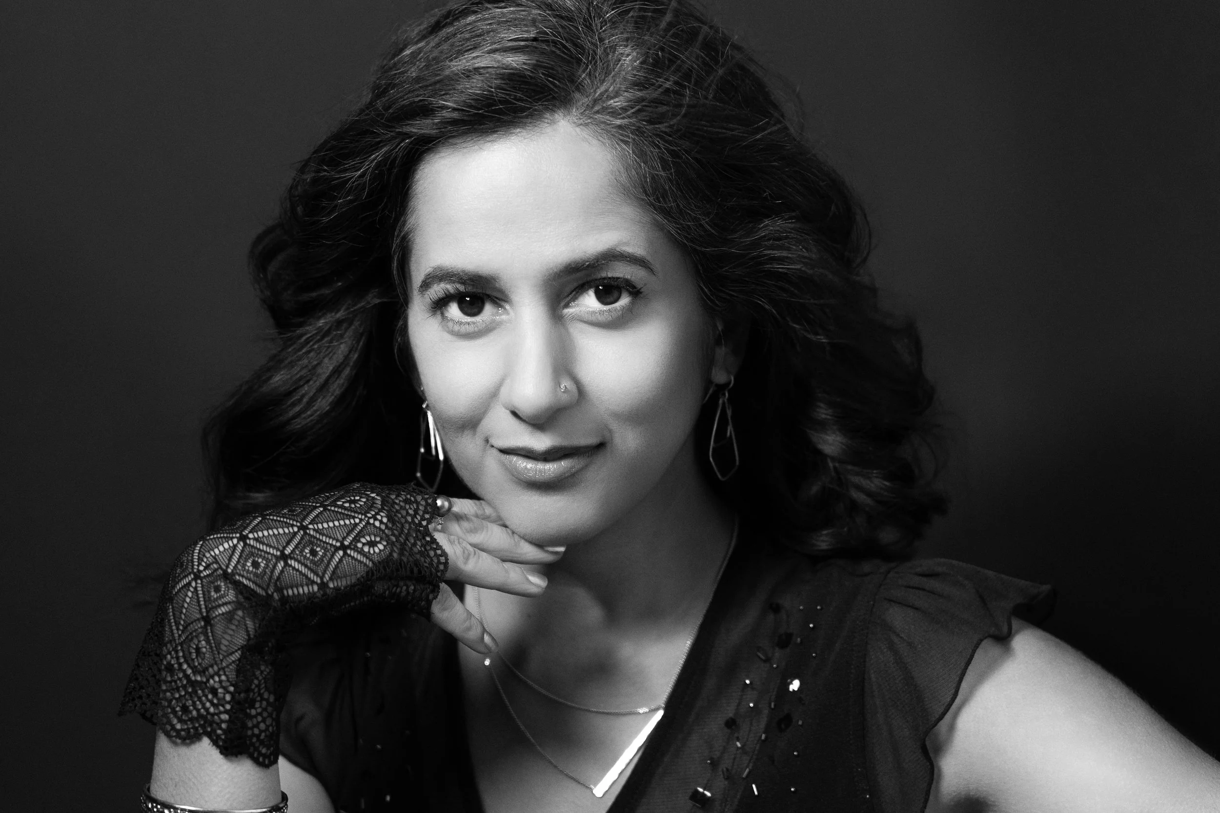 Black and white portrait of a young woman with wavy hair, wearing lace gloves, hoop earrings, and a black top with embellishments, resting her chin on her hand with a confident expression.