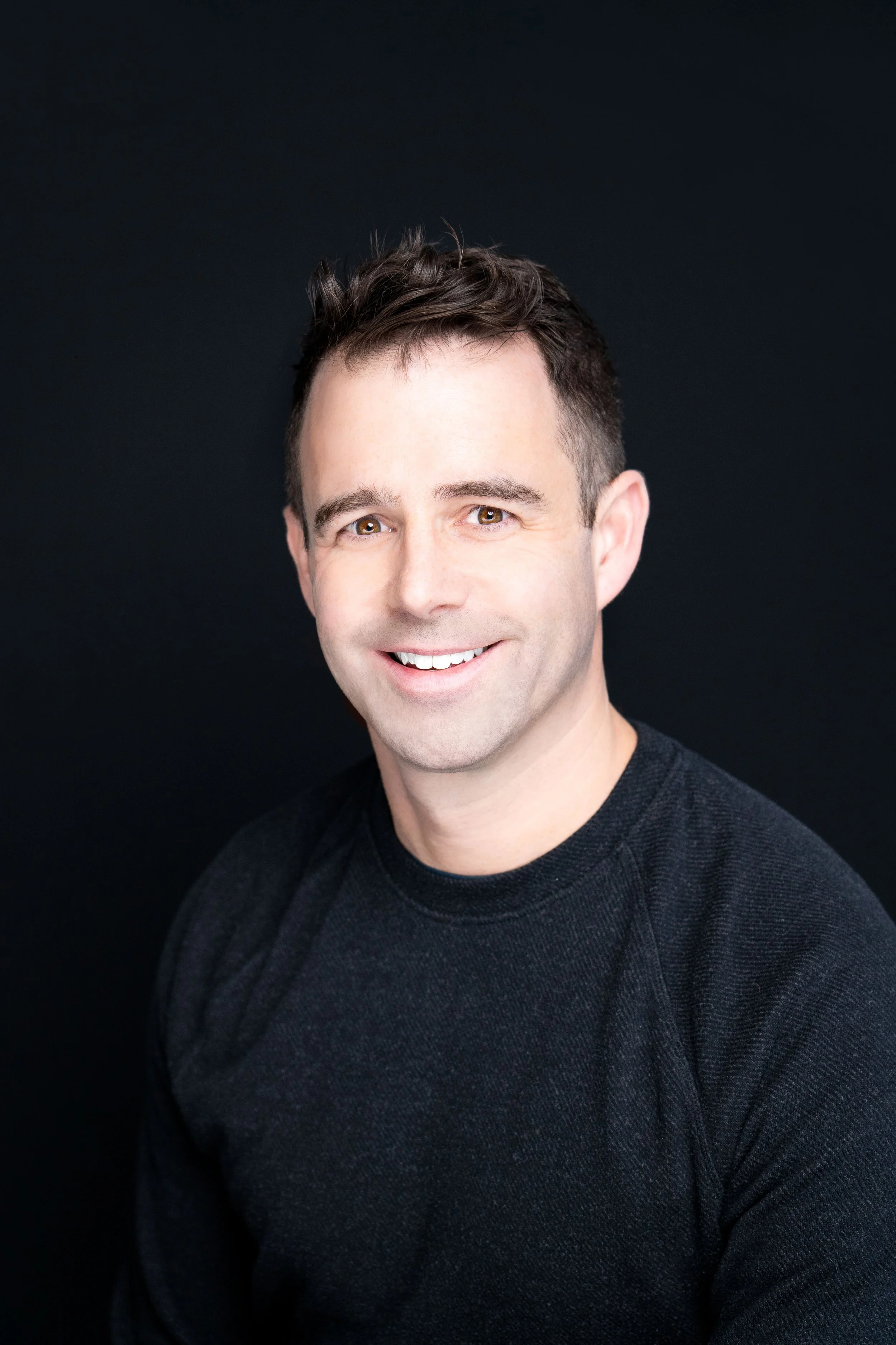 Professional headshot of a smiling man with short dark hair, wearing a black sweater, against a black background.