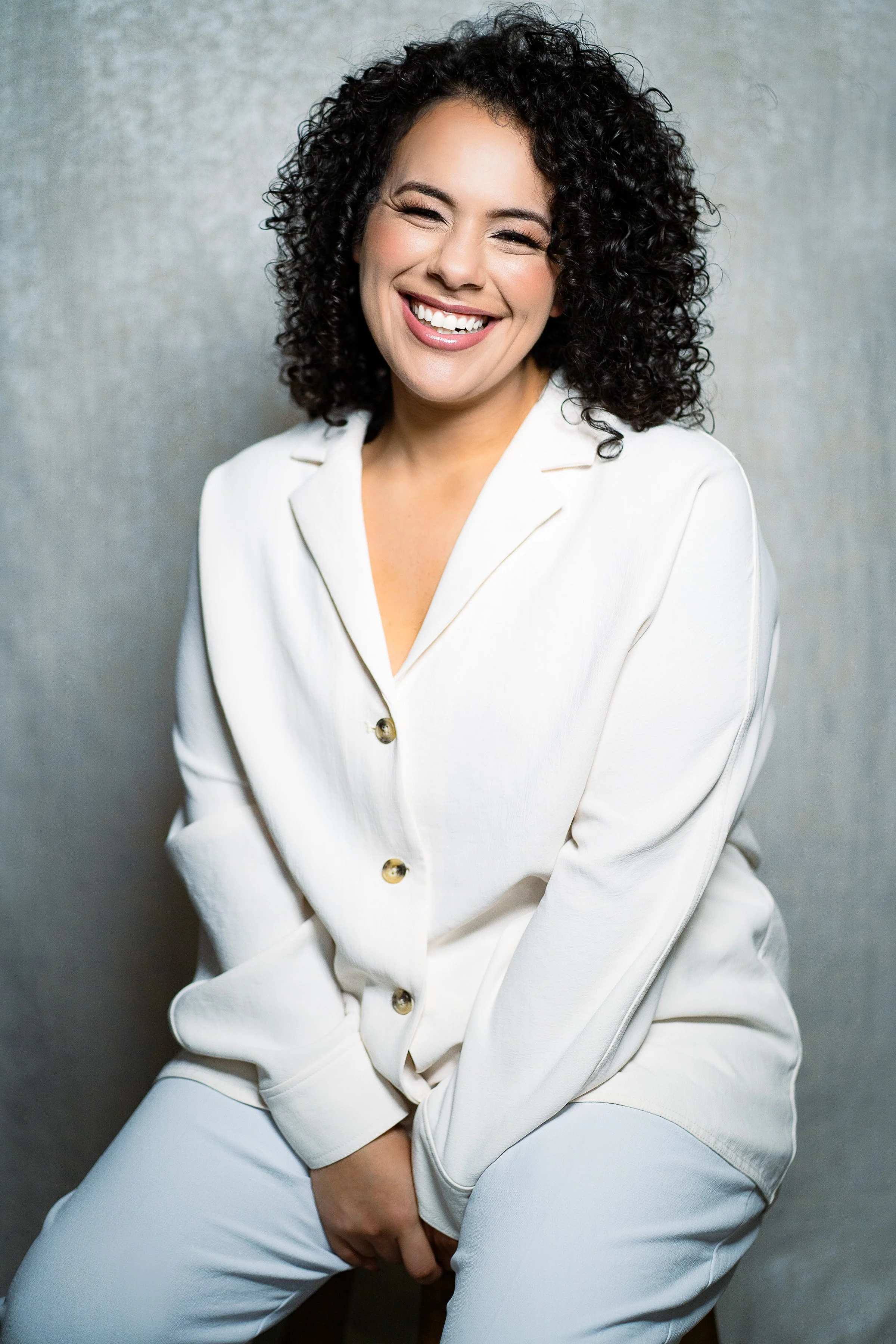A woman with curly black hair and light skin, wearing a white blazer and matching pants, is smiling and sitting against a textured gray background.