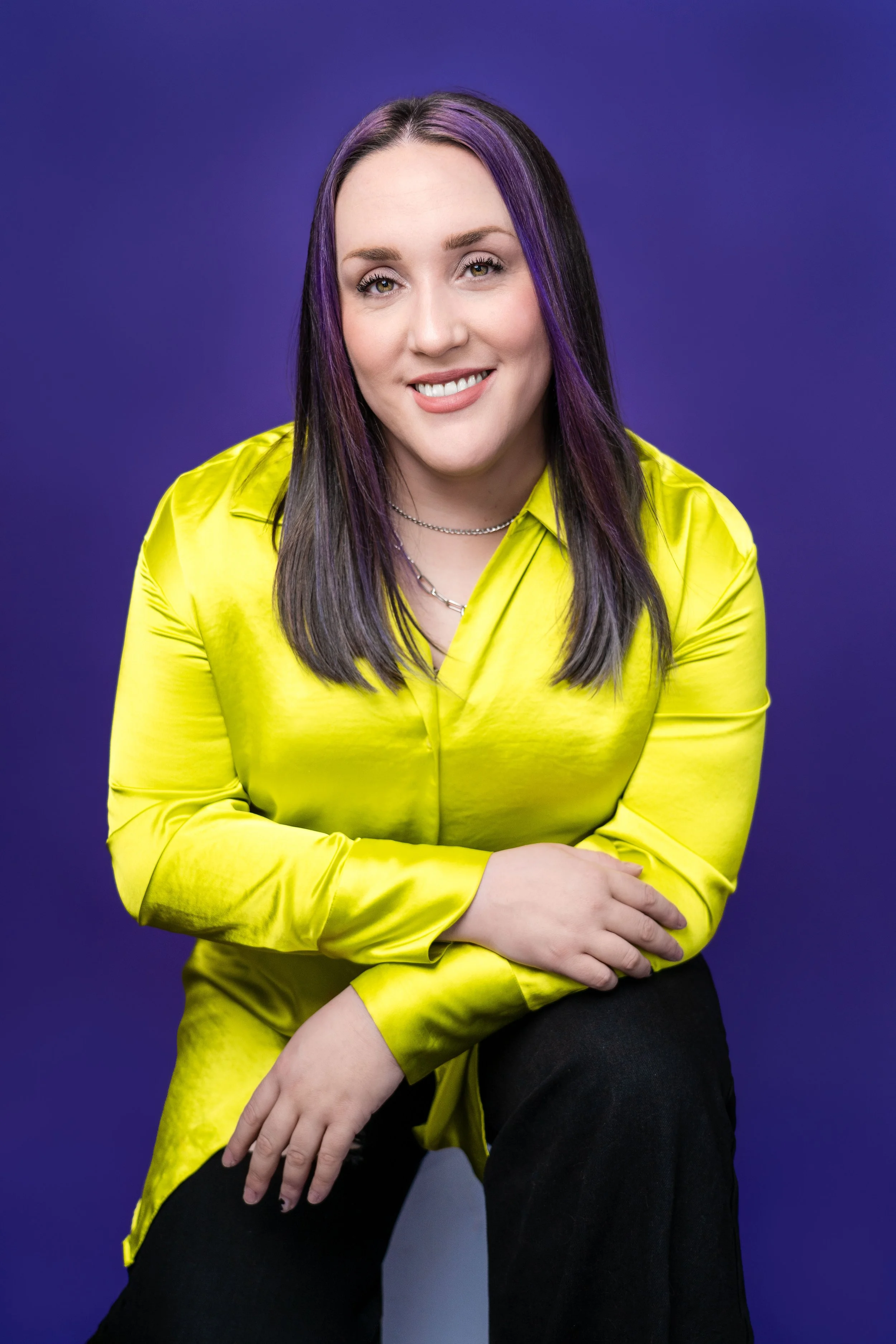 A young woman with purple and black hair, wearing a bright yellow satin shirt, smiling at the camera against a purple background.