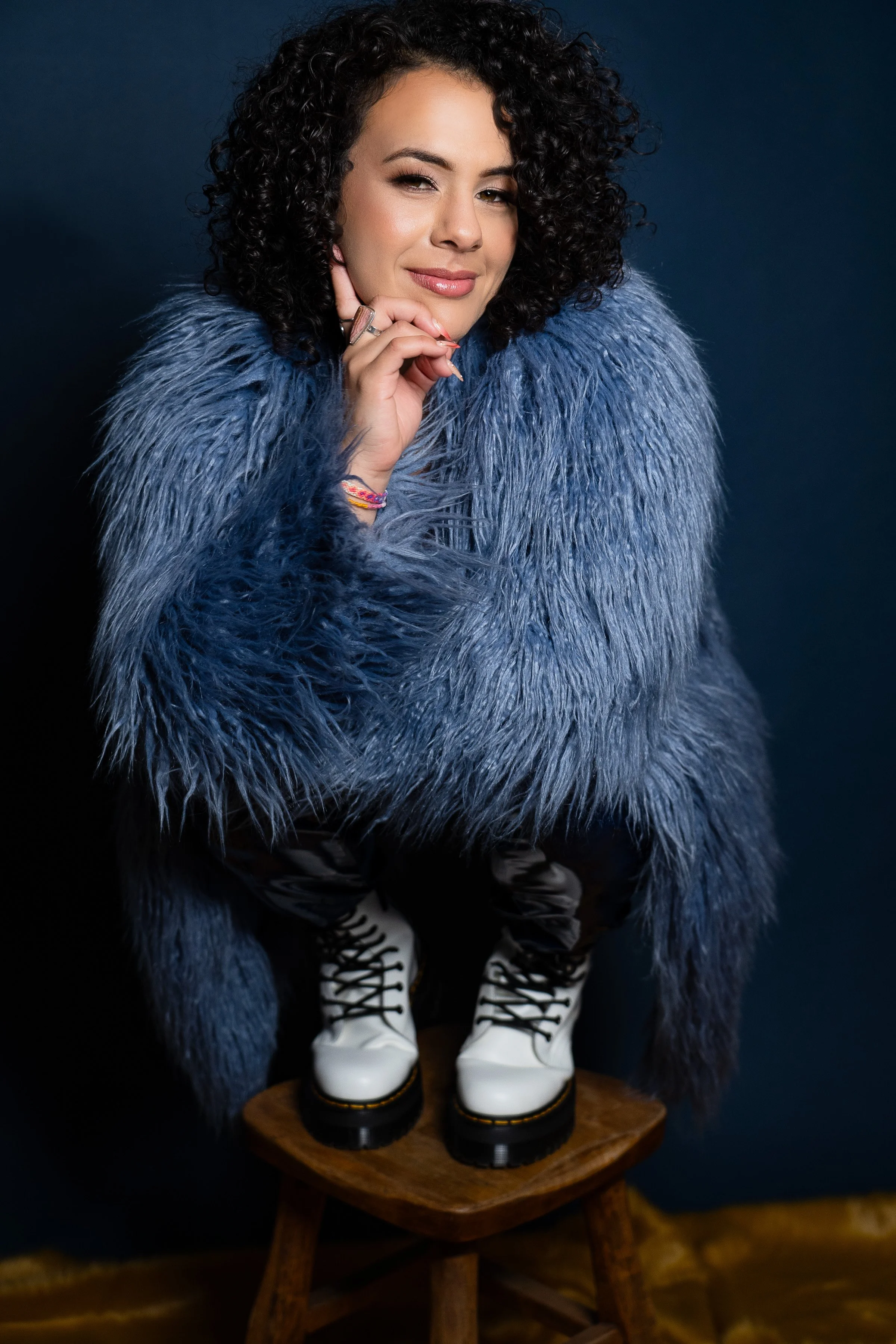 A woman with curly dark hair wearing a large, fluffy blue coat, white lace-up boots, and gold earrings, sitting on a wooden stool with her chin resting on her hand, against a dark background.