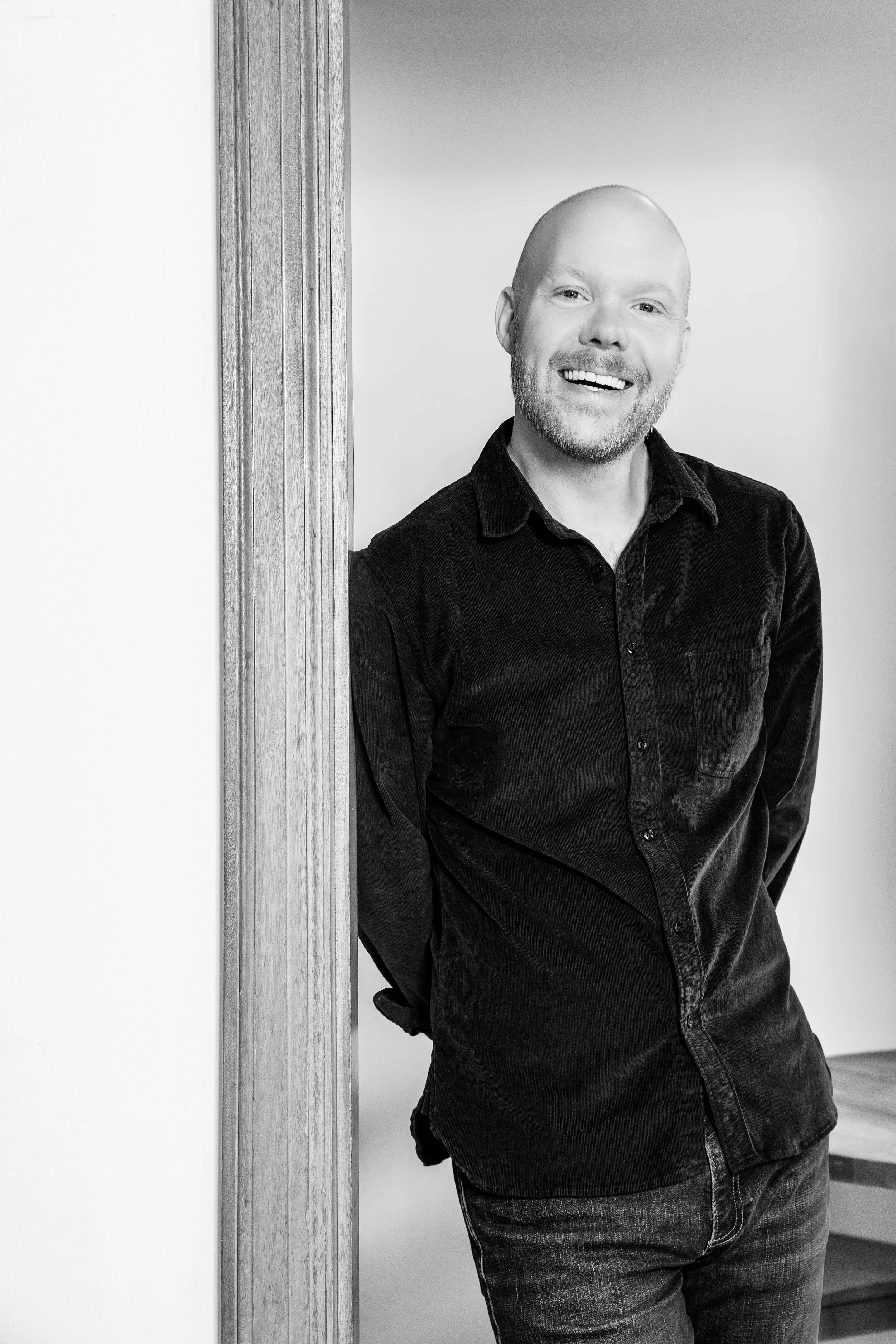 Black and white portrait of a smiling man with a beard and a shaved head, wearing a dark button-up shirt, leaning against a doorframe.