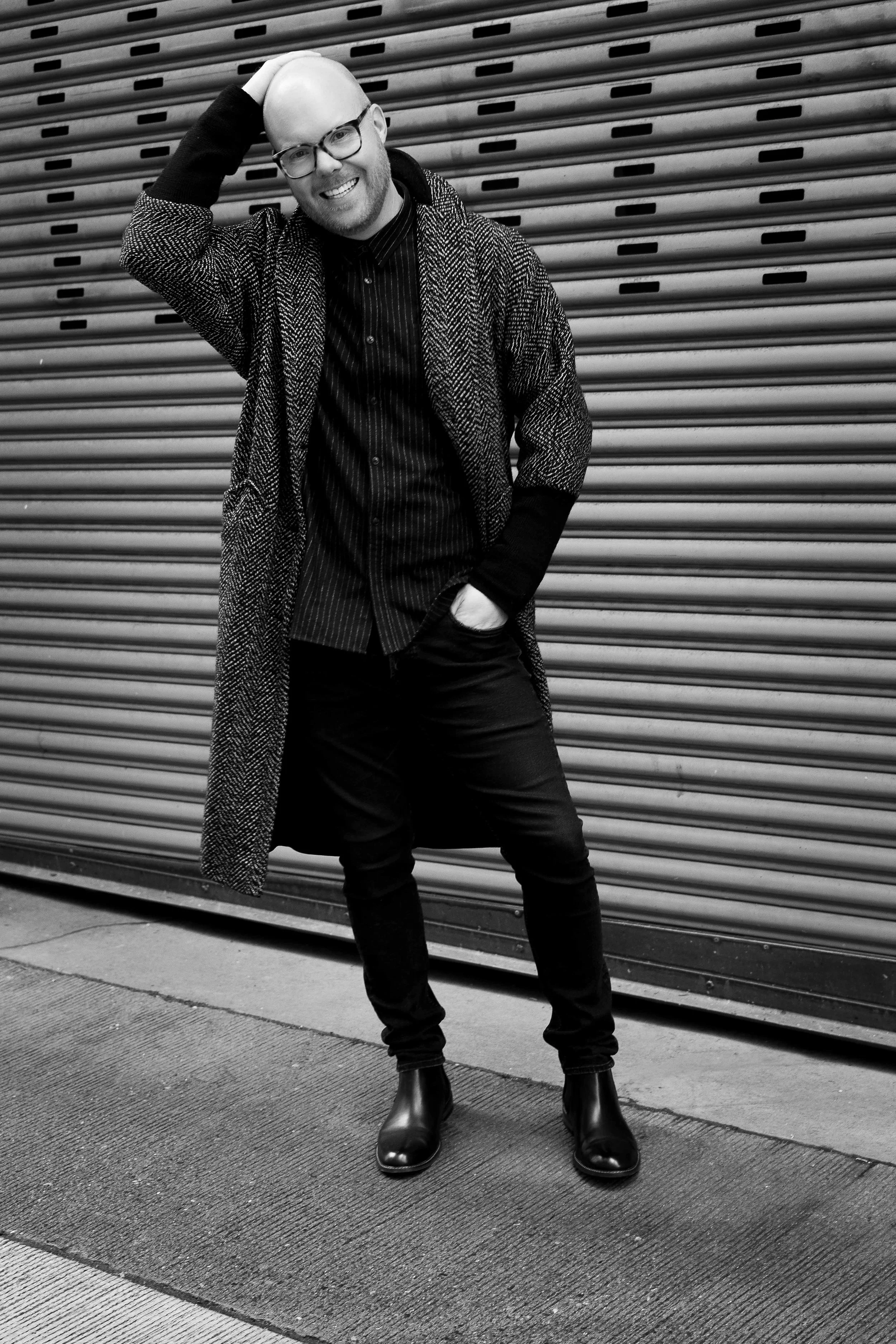 Black and white photo of a smiling man with glasses, bald head, wearing a patterned coat, striped shirt, black pants, and black boots, standing in front of horizontal metal doors, with one hand on his head and the other in his pocket.