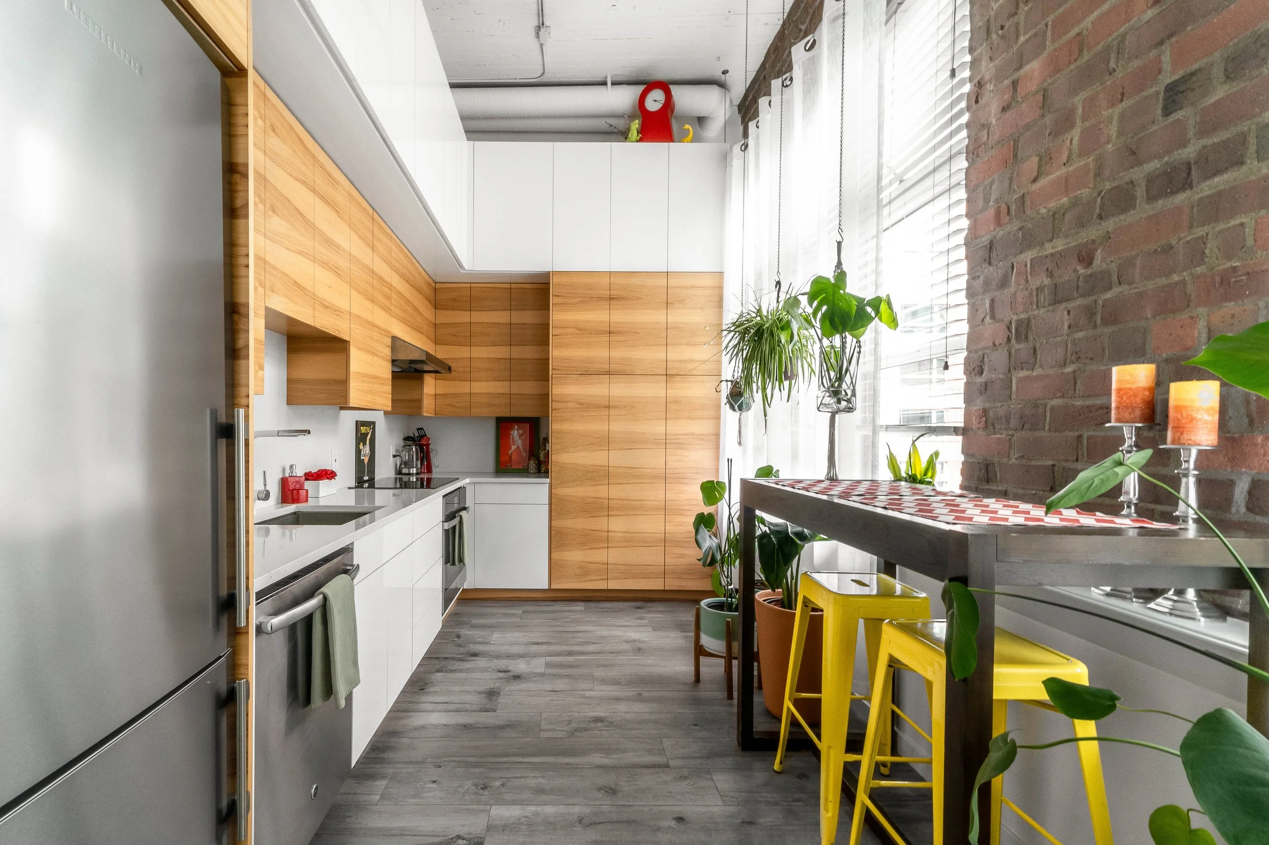 The Portrait Playdate Seattle studio's modern-kitchen-slash-hair-and-makeup-station with wood cabinets, white countertops, stainless steel appliances, hanging plants near a large window, brick wall, and yellow chairs at a small dining table.