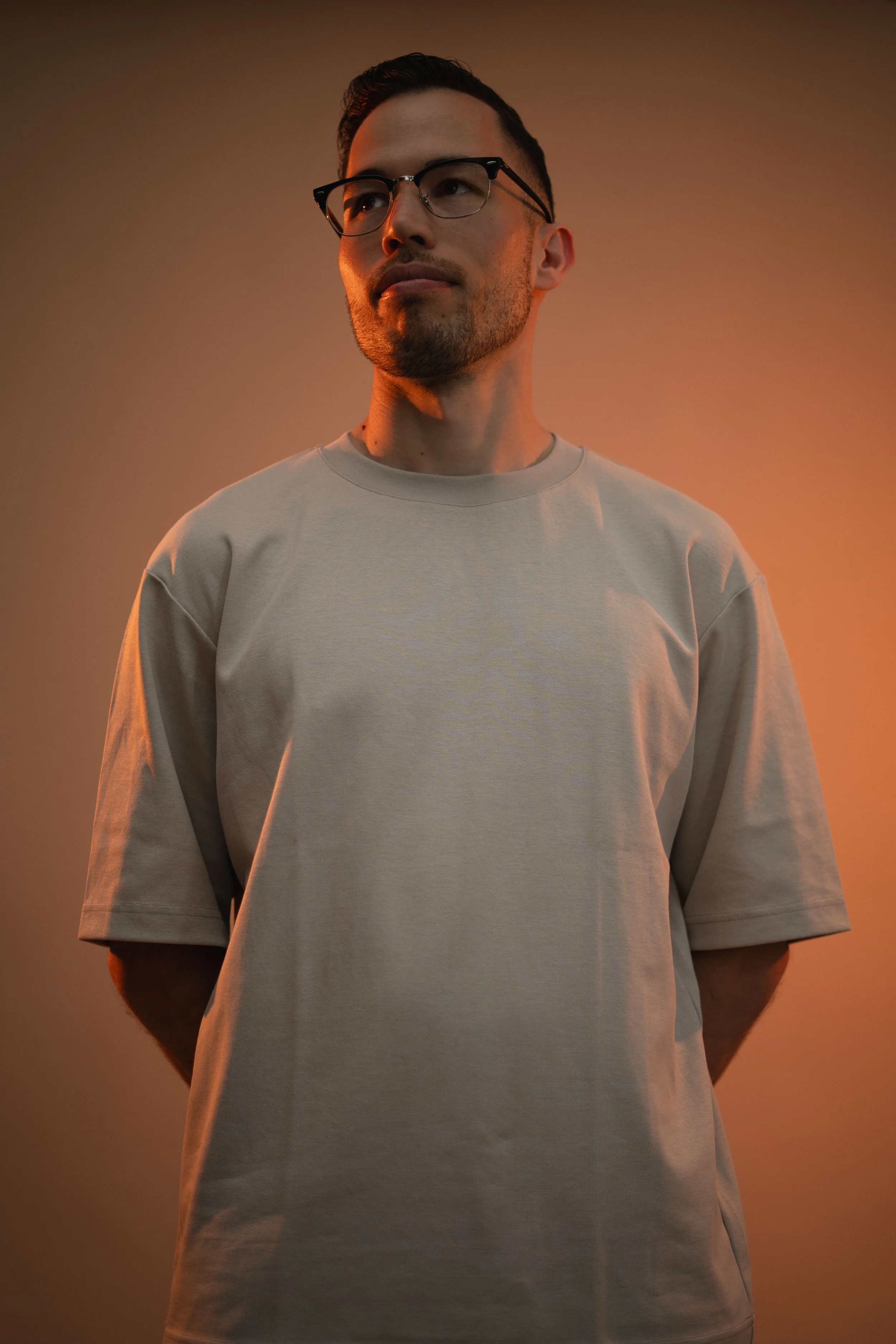 A man with glasses and a beard, wearing a beige oversized t-shirt, stands against a plain background illuminated with warm orange lighting.