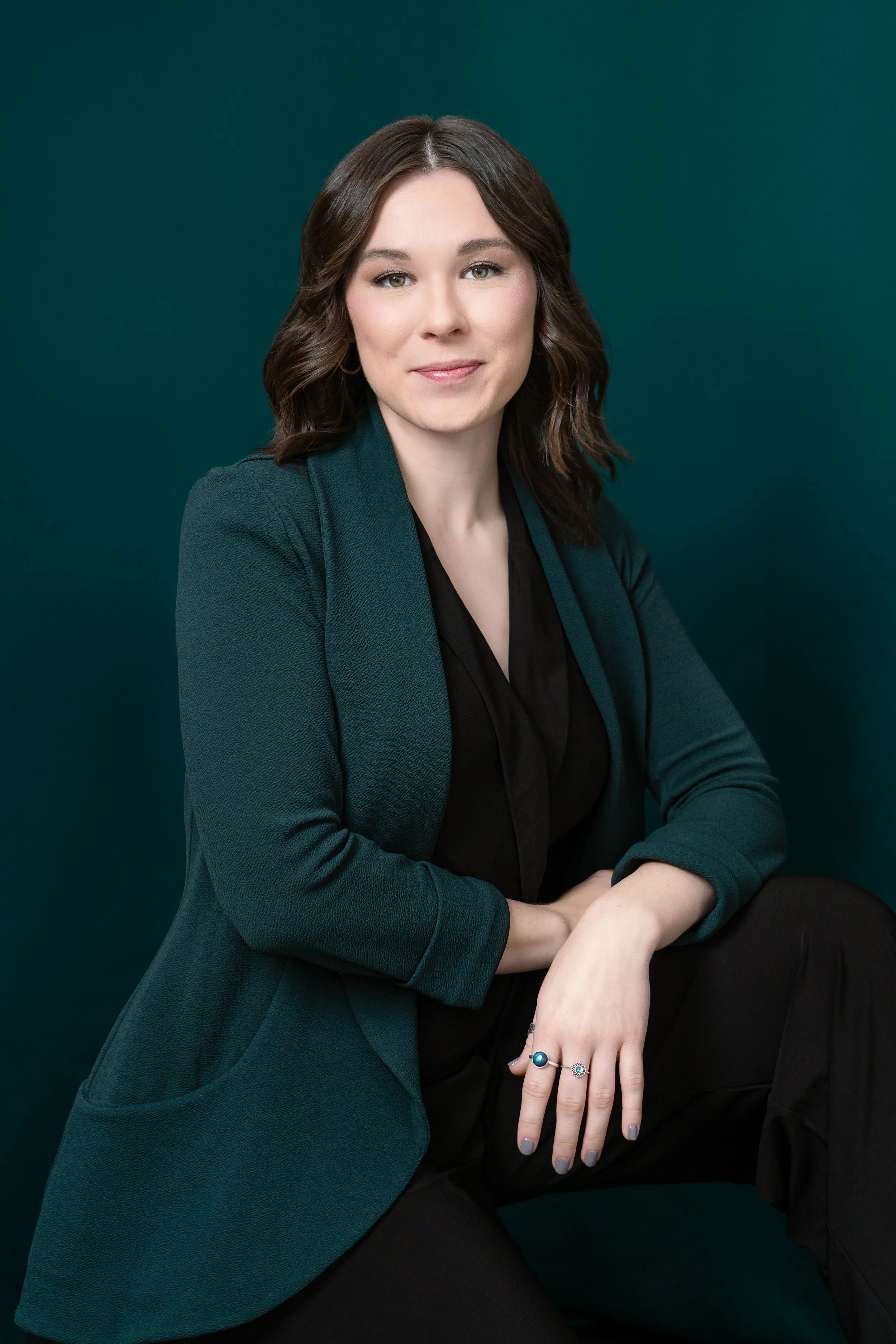 Professional woman with brown wavy hair, wearing a dark teal blazer and black top, sitting against a solid dark green background.