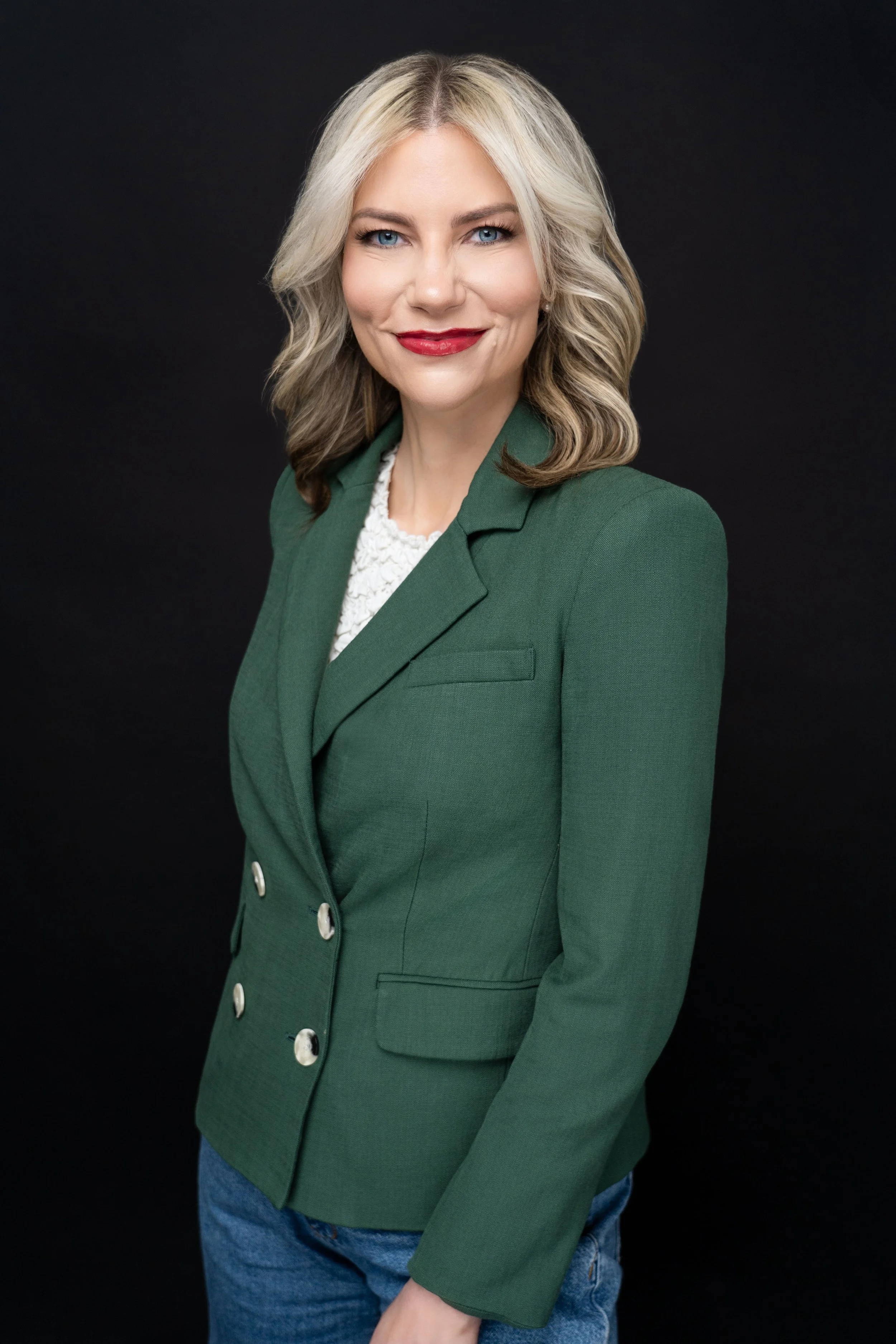 A woman with shoulder-length blonde hair, wearing a green blazer, white top, and blue jeans, smiling against a dark background.