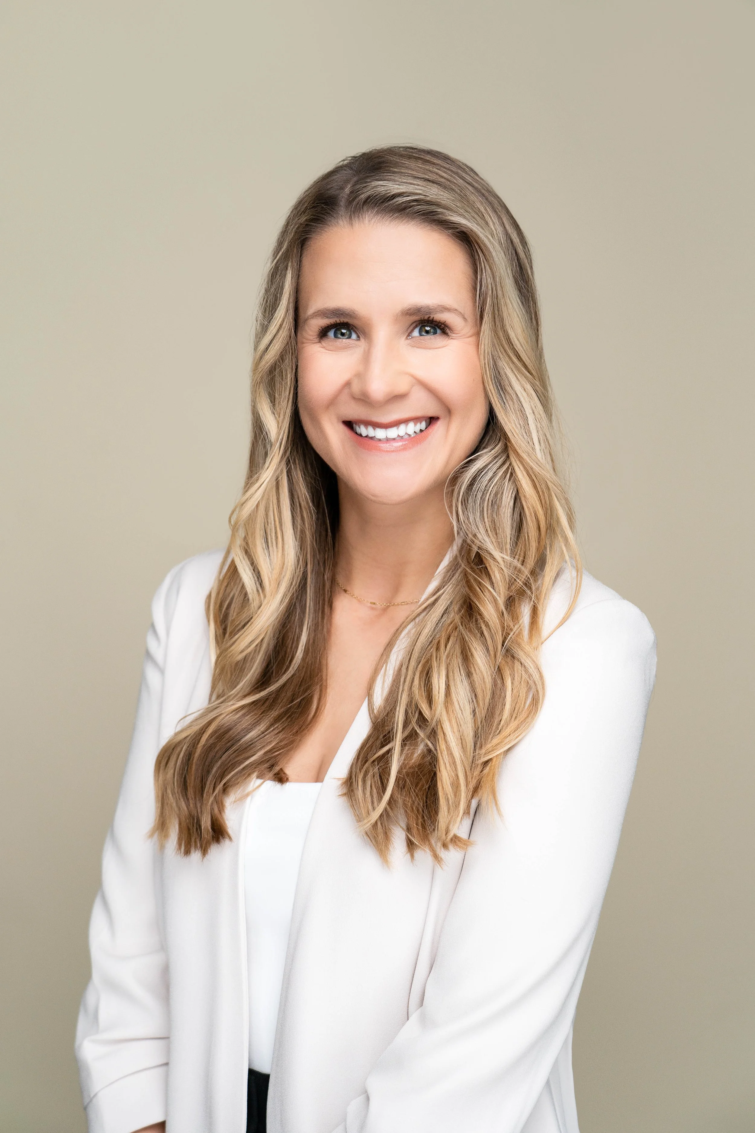 Portrait of a smiling woman with long wavy blonde hair, wearing a white blazer and top, against a plain light background.