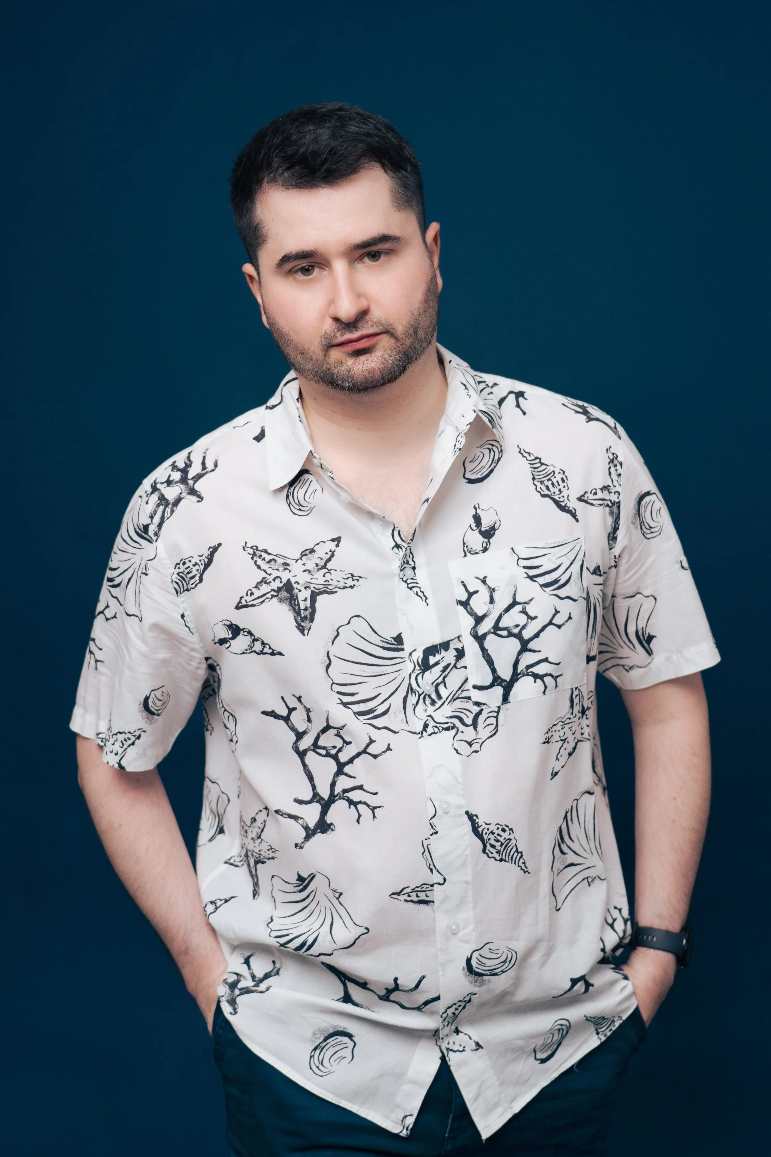 A man with short dark hair, beard, and mustache wearing a white short-sleeve shirt with black seashell, starfish, and coral patterns, standing against a solid dark blue background with his hands in his pockets.
