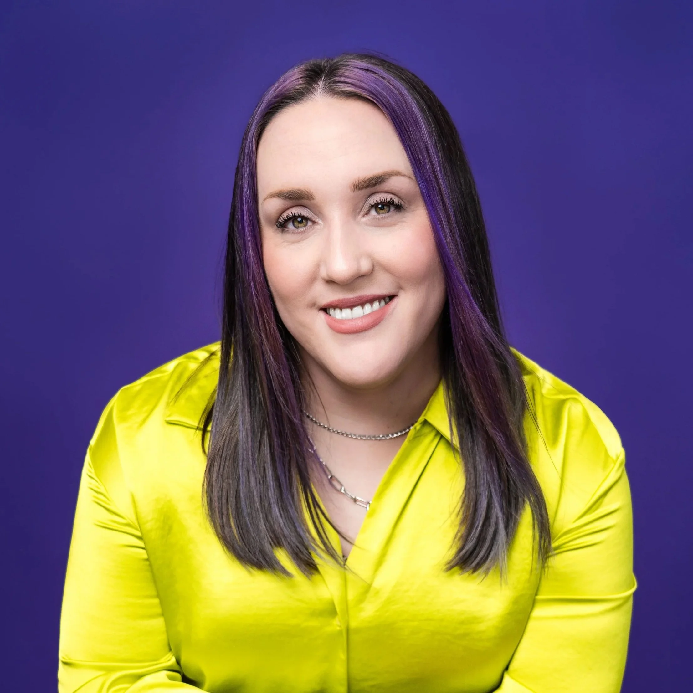 Headshot of a woman with purple hair and a chartreuse sikly shirt against a dark purple background
