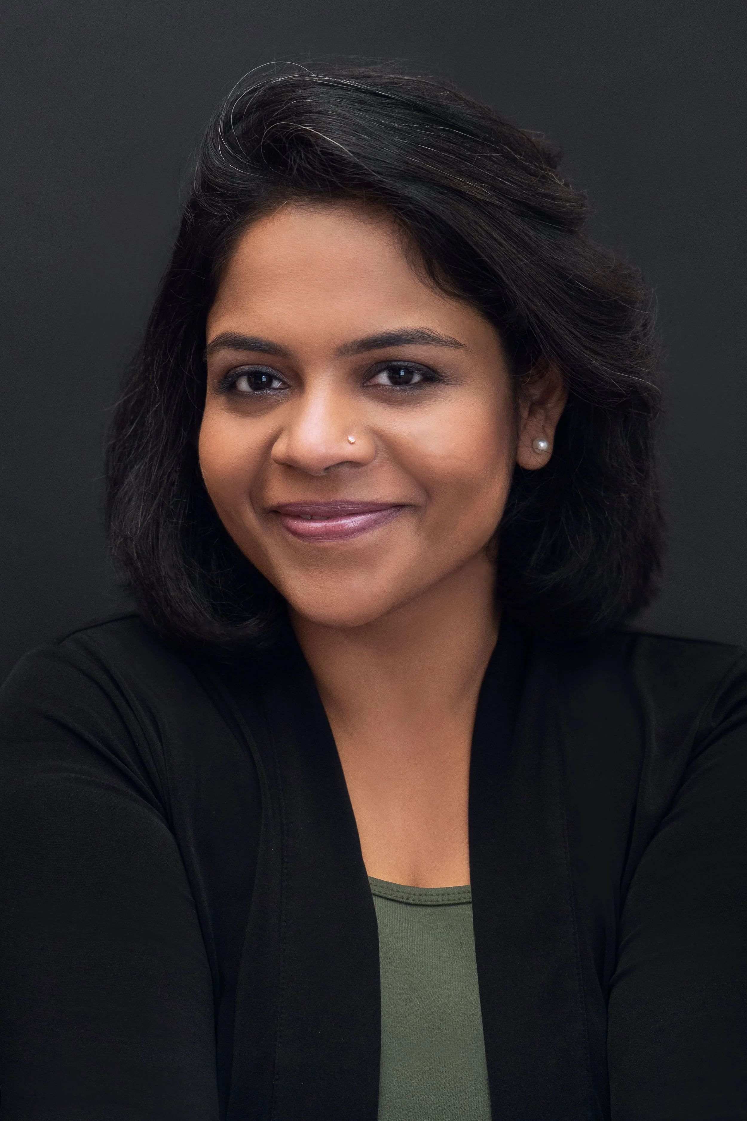 Headshot of a woman with medium-length black hair, wearing a black jacket over a green top, smiling against a dark background.