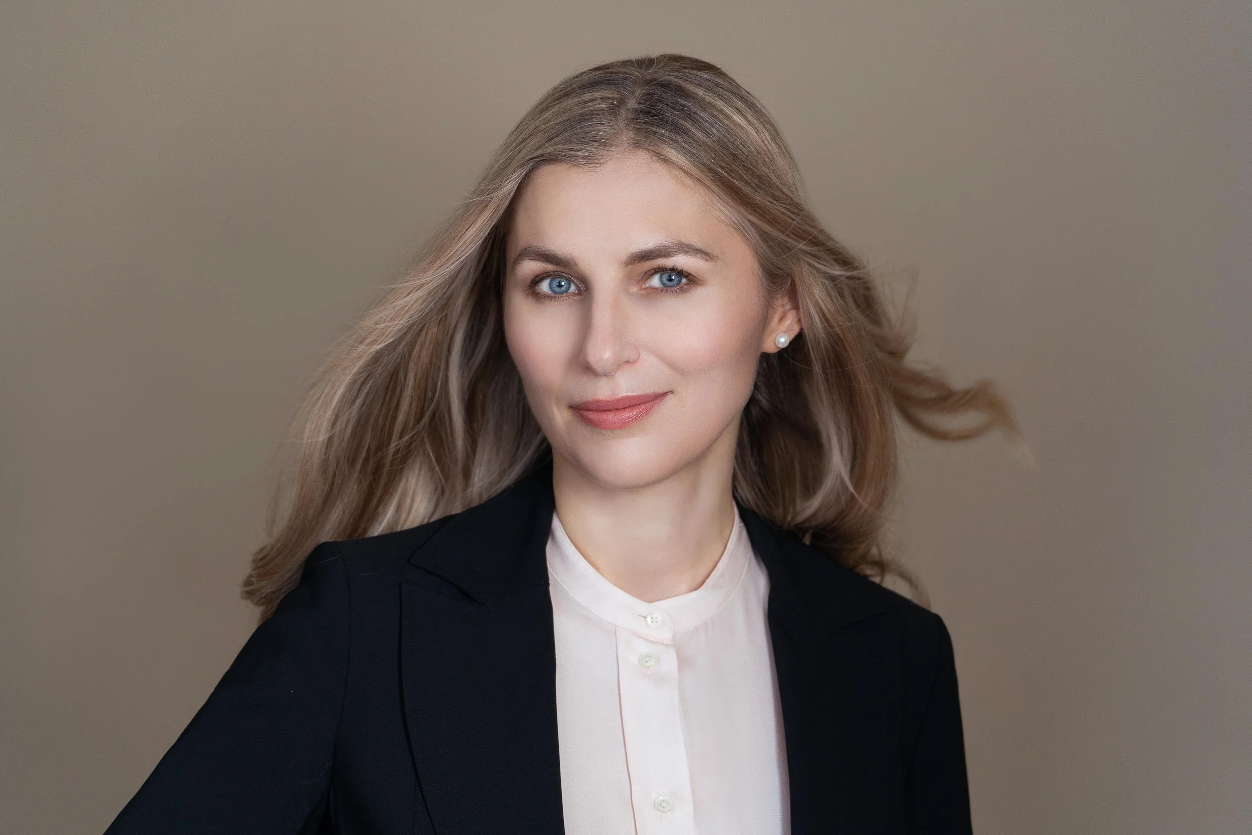 Portrait of a woman with blonde hair, blue eyes, wearing a black blazer and white blouse, smiling softly against a neutral background.