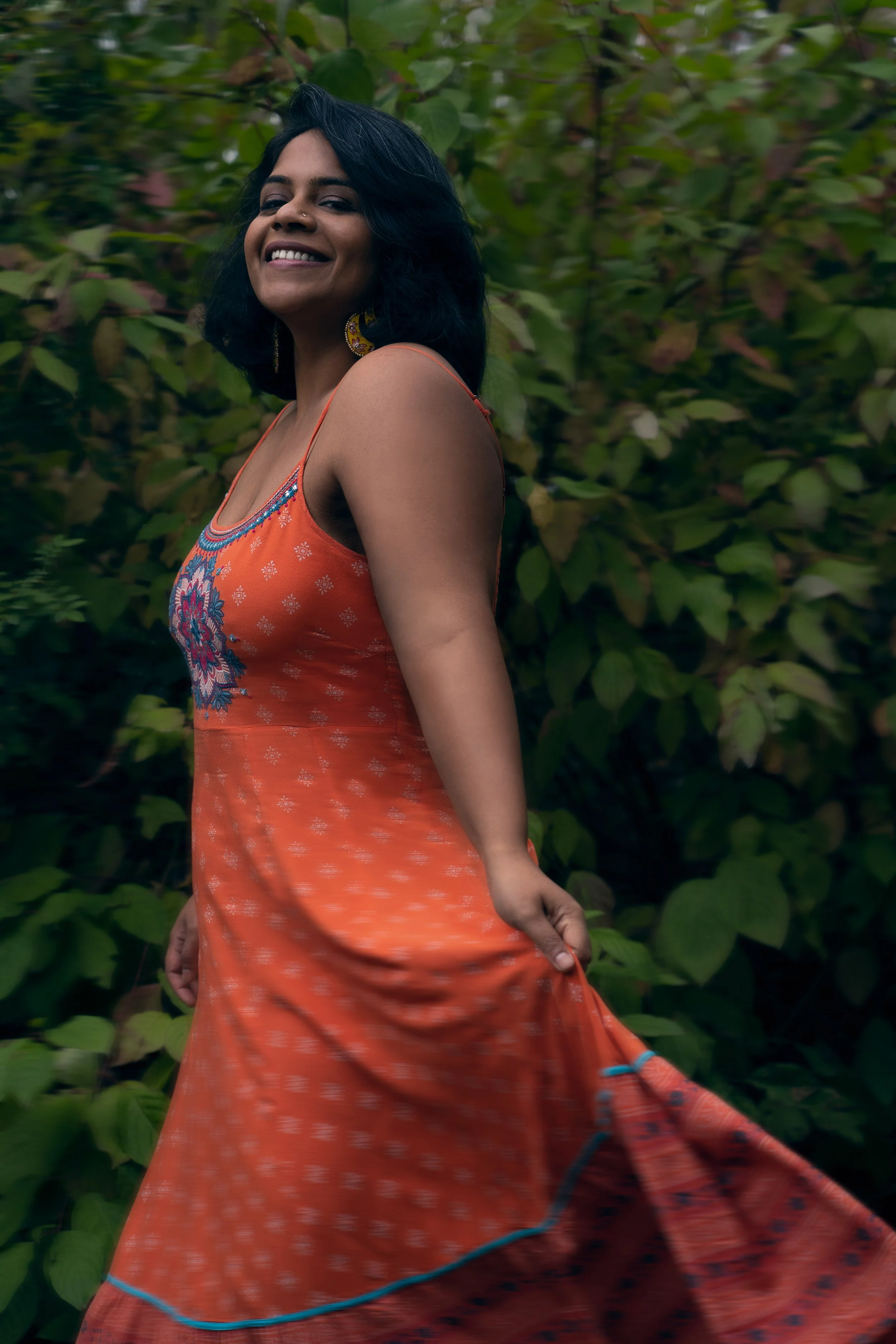 A woman with dark hair smiling, wearing an orange dress, standing outdoors near green foliage.